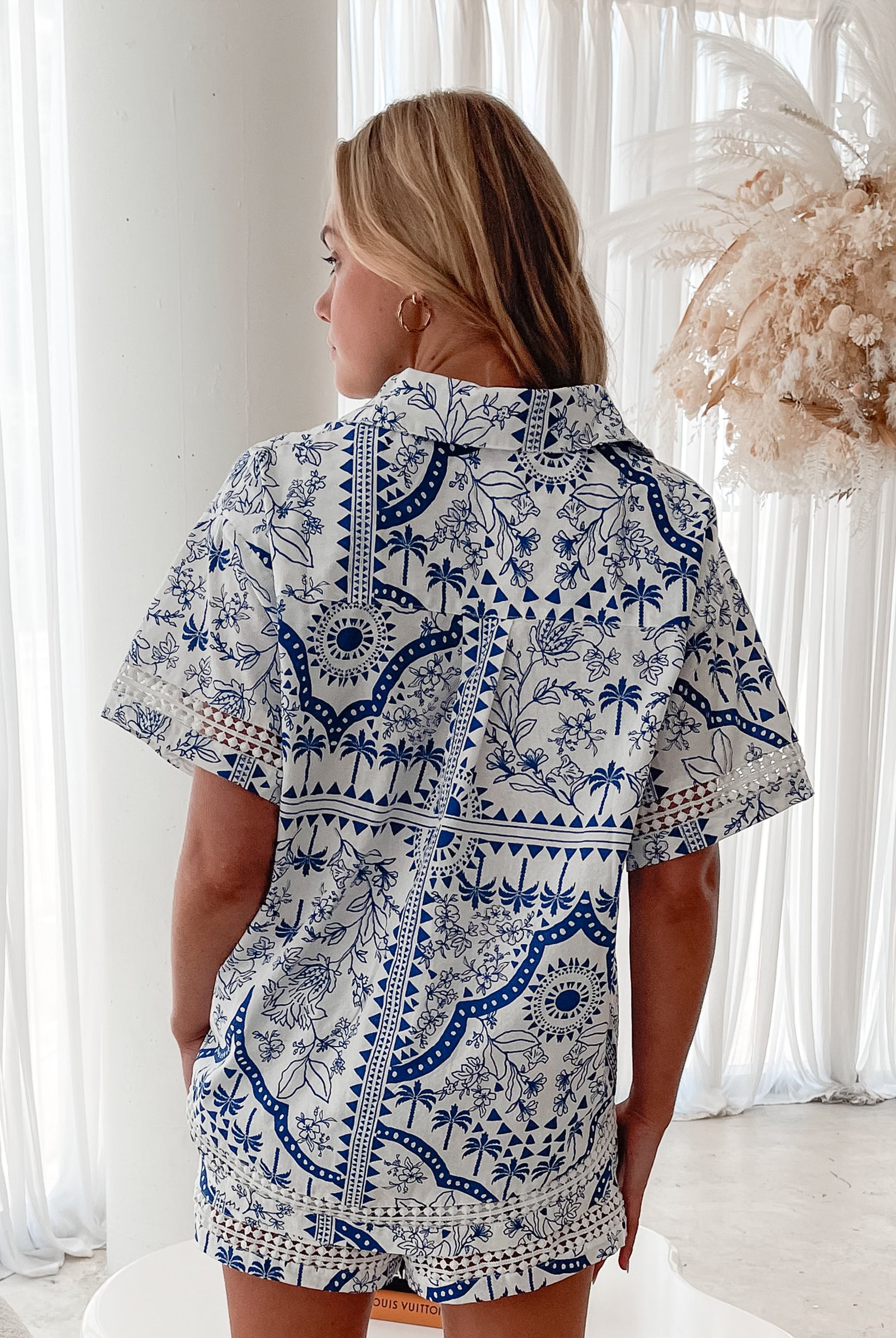A woman with blonde, wavy hair stands in a bright, modern room with floor-to-ceiling sheer white curtains and neutral-toned decor. She is wearing a coordinated short-sleeve button-up shirt and matching shorts set featuring a blue and white floral and geometric print. She has a relaxed expression with one hand in her pocket, exuding a stylish and effortless vibe.