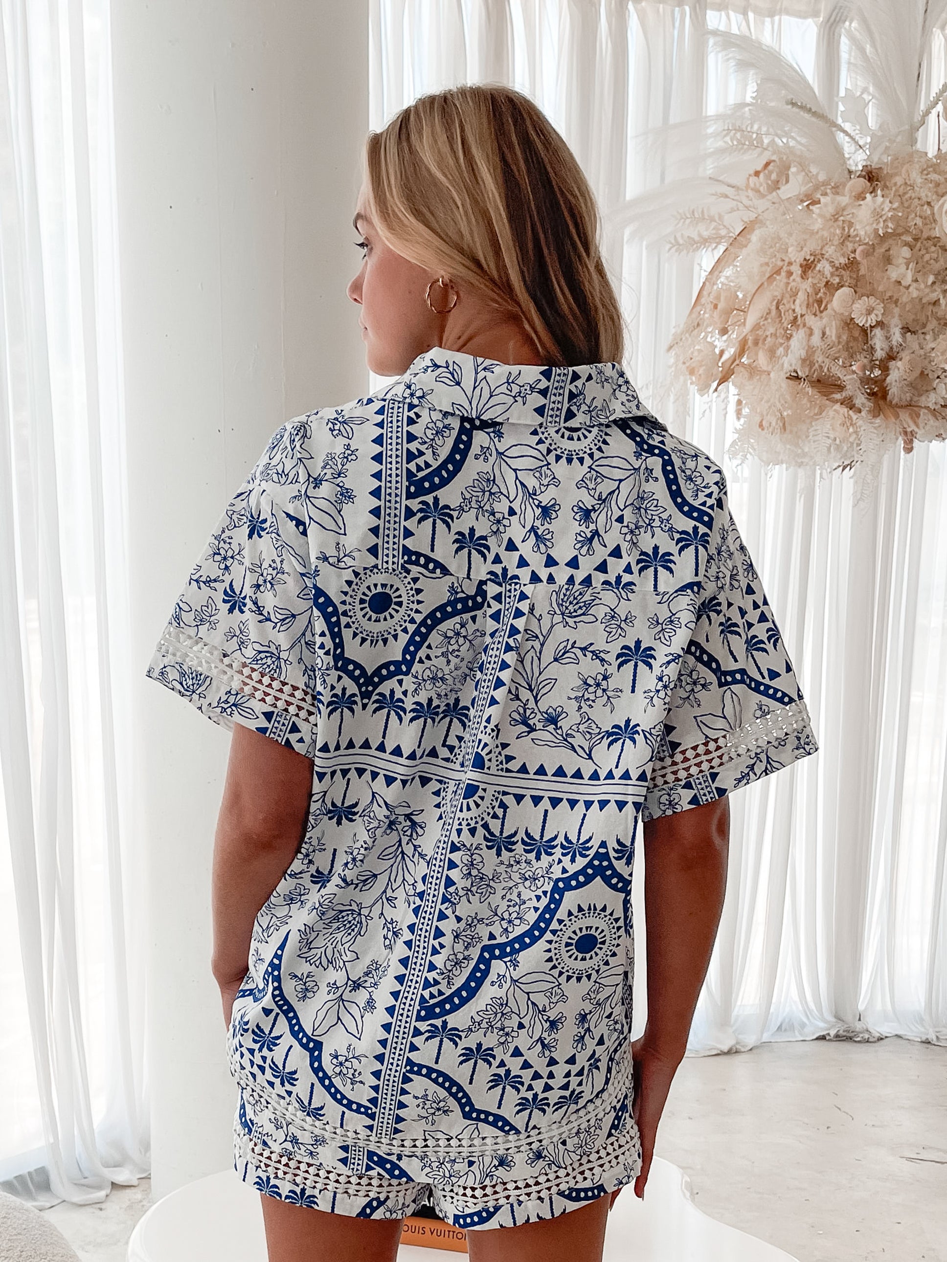 A woman with blonde, wavy hair stands in a bright, modern room with floor-to-ceiling sheer white curtains and neutral-toned decor. She is wearing a coordinated short-sleeve button-up shirt and matching shorts set featuring a blue and white floral and geometric print. She has a relaxed expression with one hand in her pocket, exuding a stylish and effortless vibe.