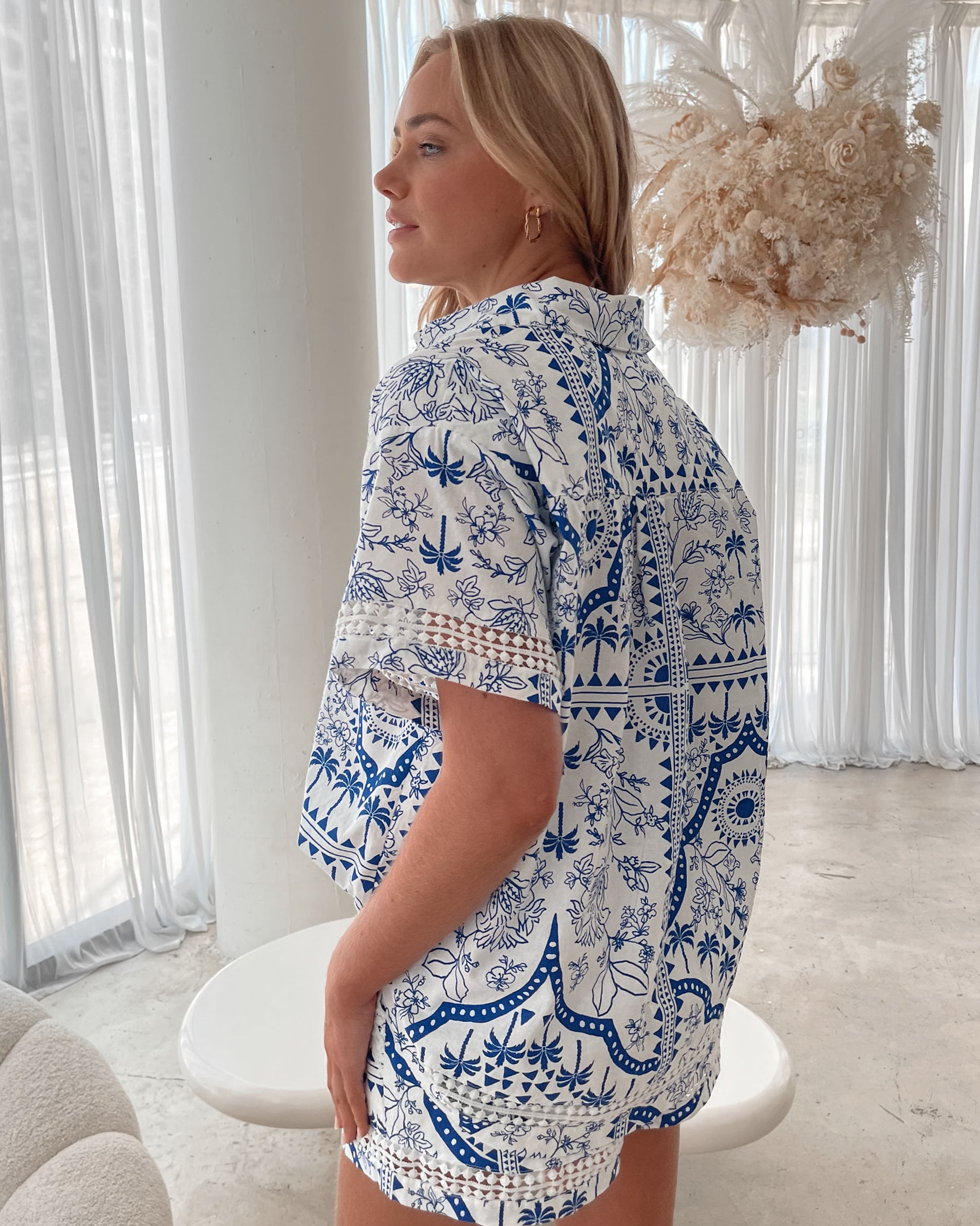 A woman with blonde, wavy hair stands in a bright, modern room with floor-to-ceiling sheer white curtains and neutral-toned decor. She is wearing a coordinated short-sleeve button-up shirt and matching shorts set featuring a blue and white floral and geometric print. She has a relaxed expression with one hand in her pocket, exuding a stylish and effortless vibe.