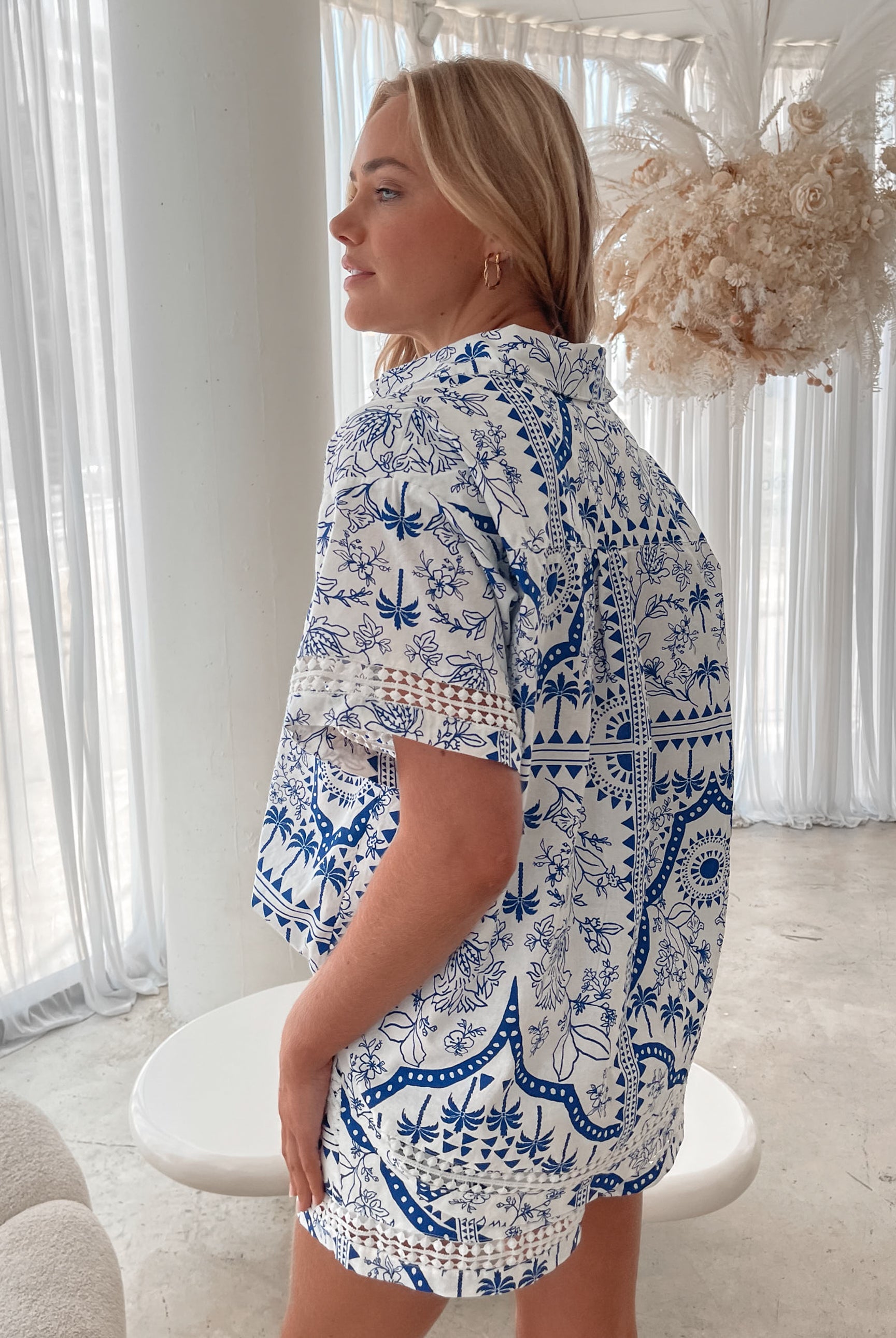A woman with blonde, wavy hair stands in a bright, modern room with floor-to-ceiling sheer white curtains and neutral-toned decor. She is wearing a coordinated short-sleeve button-up shirt and matching shorts set featuring a blue and white floral and geometric print. She has a relaxed expression with one hand in her pocket, exuding a stylish and effortless vibe.
