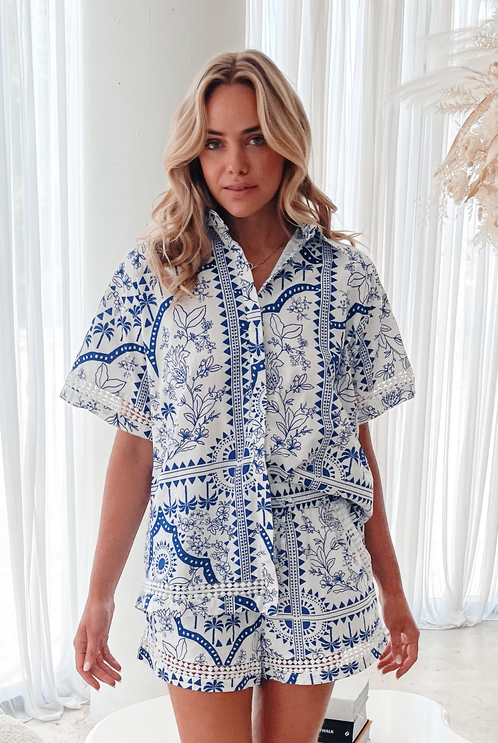 A woman with blonde, wavy hair stands in a bright, modern room with floor-to-ceiling sheer white curtains and neutral-toned decor. She is wearing a coordinated short-sleeve button-up shirt and matching shorts set featuring a blue and white floral and geometric print. She has a relaxed expression with one hand in her pocket, exuding a stylish and effortless vibe.