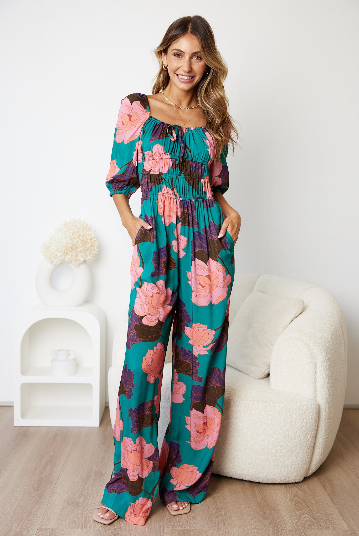 A woman with long, wavy blonde hair sits on the edge of a cream-colored seat, wearing a vibrant teal jumpsuit adorned with large pink and purple floral prints. The jumpsuit features a ruched bodice with a tie detail and puffed three-quarter sleeves. She has a relaxed, confident expression, with one hand in her hair, set against a simple white wall background.