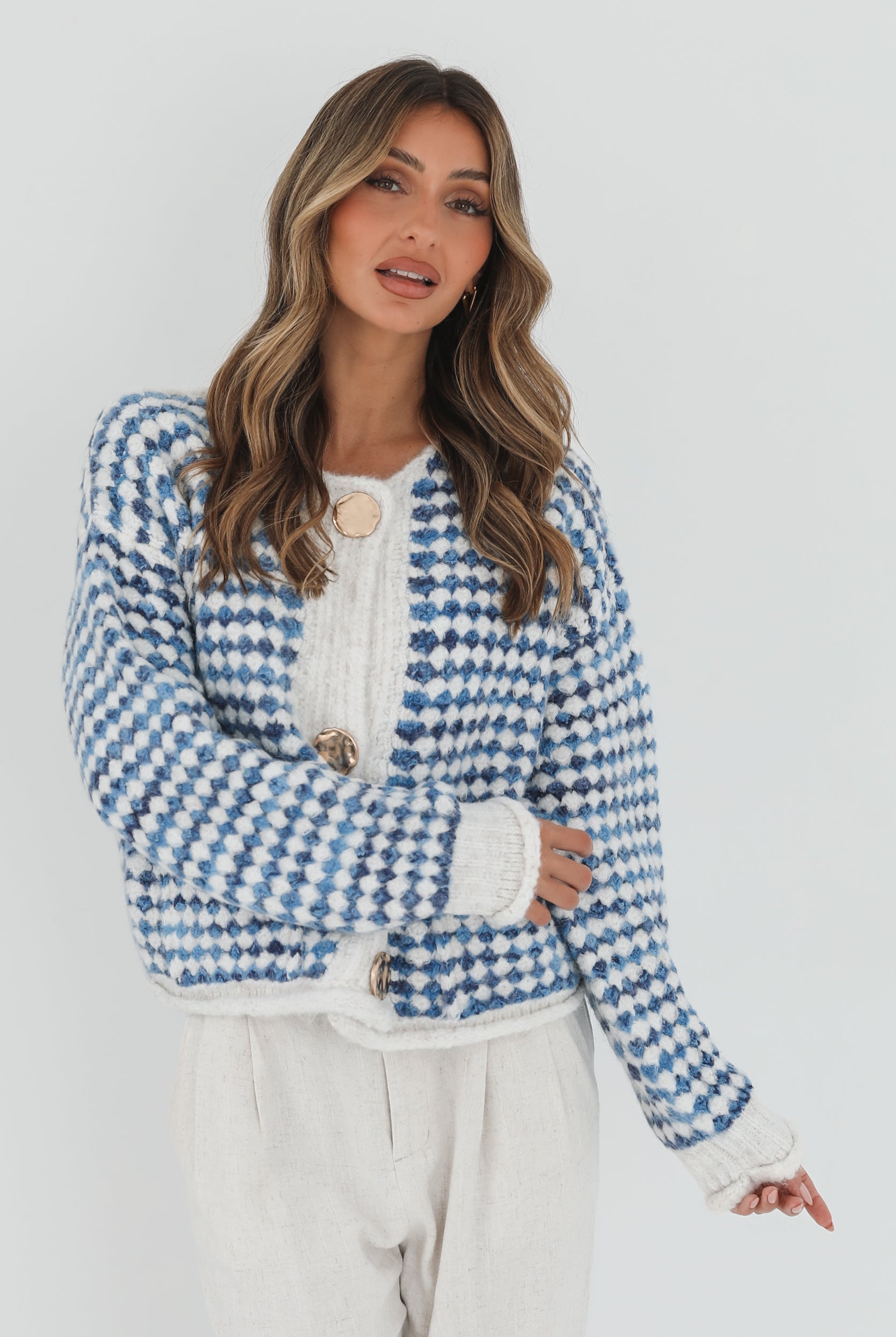 A woman with long, wavy brown hair stands against a plain white background, wearing a cozy blue and white checkered cardigan with oversized gold buttons and front pockets. She pairs it with light cream-colored pants and has her hands casually tucked into her pockets, giving a confident and composed look.