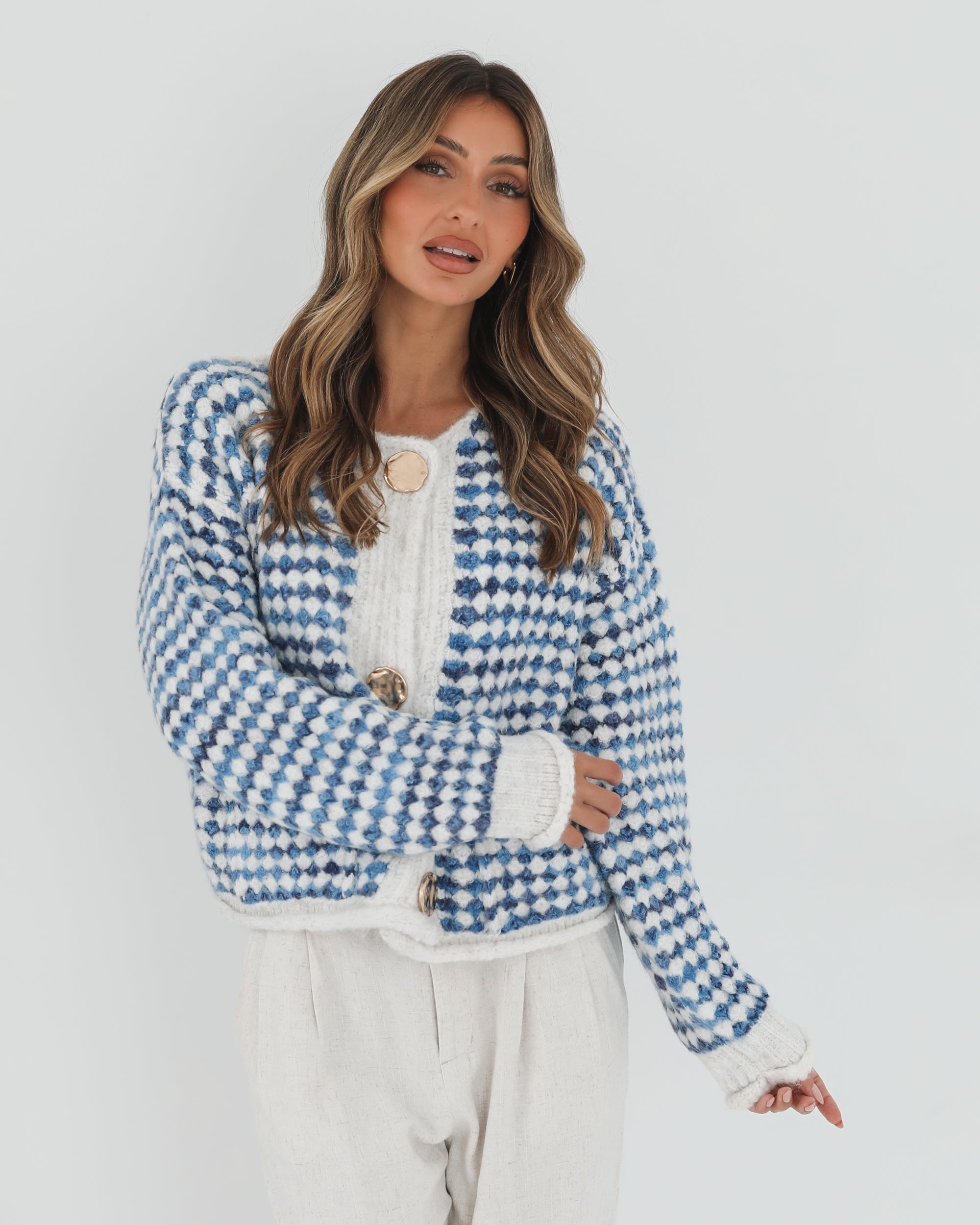 A woman with long, wavy brown hair stands against a plain white background, wearing a cozy blue and white checkered cardigan with oversized gold buttons and front pockets. She pairs it with light cream-colored pants and has her hands casually tucked into her pockets, giving a confident and composed look.