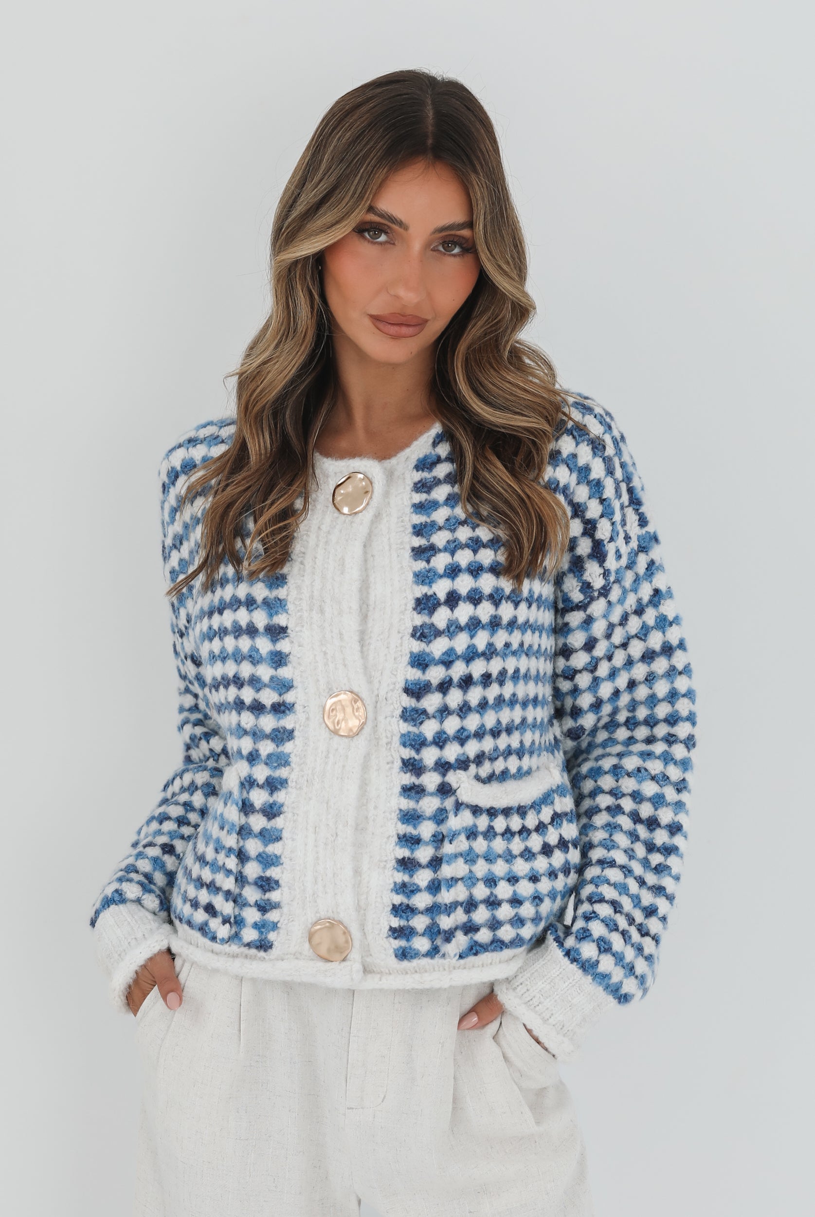 A woman with long, wavy brown hair stands against a plain white background, wearing a cozy blue and white checkered cardigan with oversized gold buttons and front pockets. She pairs it with light cream-colored pants and has her hands casually tucked into her pockets, giving a confident and composed look.