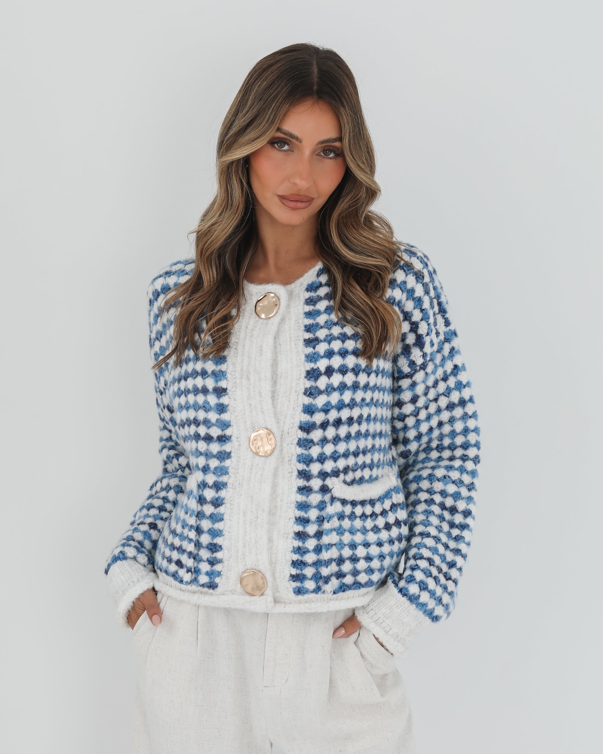 A woman with long, wavy brown hair stands against a plain white background, wearing a cozy blue and white checkered cardigan with oversized gold buttons and front pockets. She pairs it with light cream-colored pants and has her hands casually tucked into her pockets, giving a confident and composed look.
