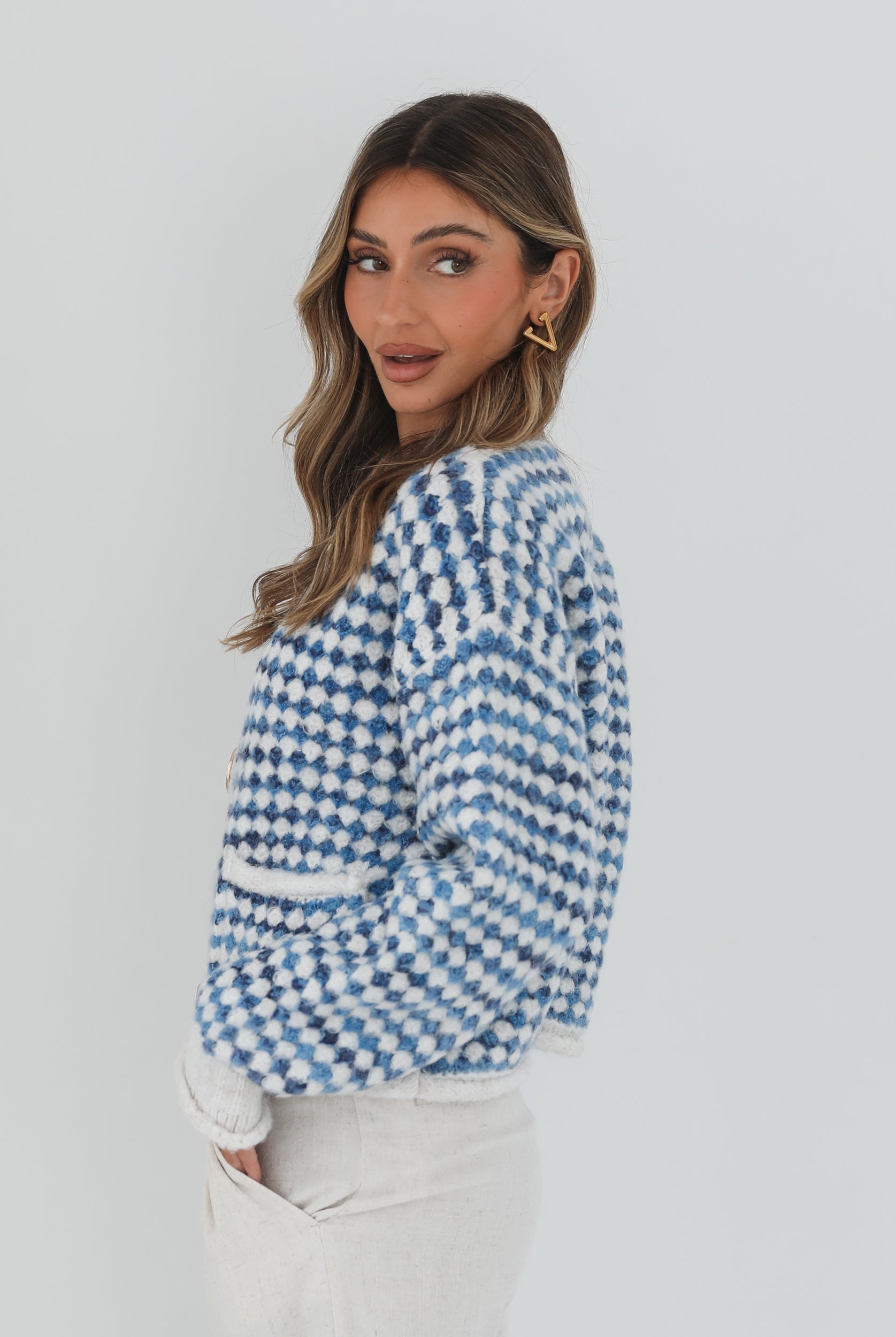 A woman with long, wavy brown hair stands against a plain white background, wearing a cozy blue and white checkered cardigan with oversized gold buttons and front pockets. She pairs it with light cream-colored pants and has her hands casually tucked into her pockets, giving a confident and composed look.