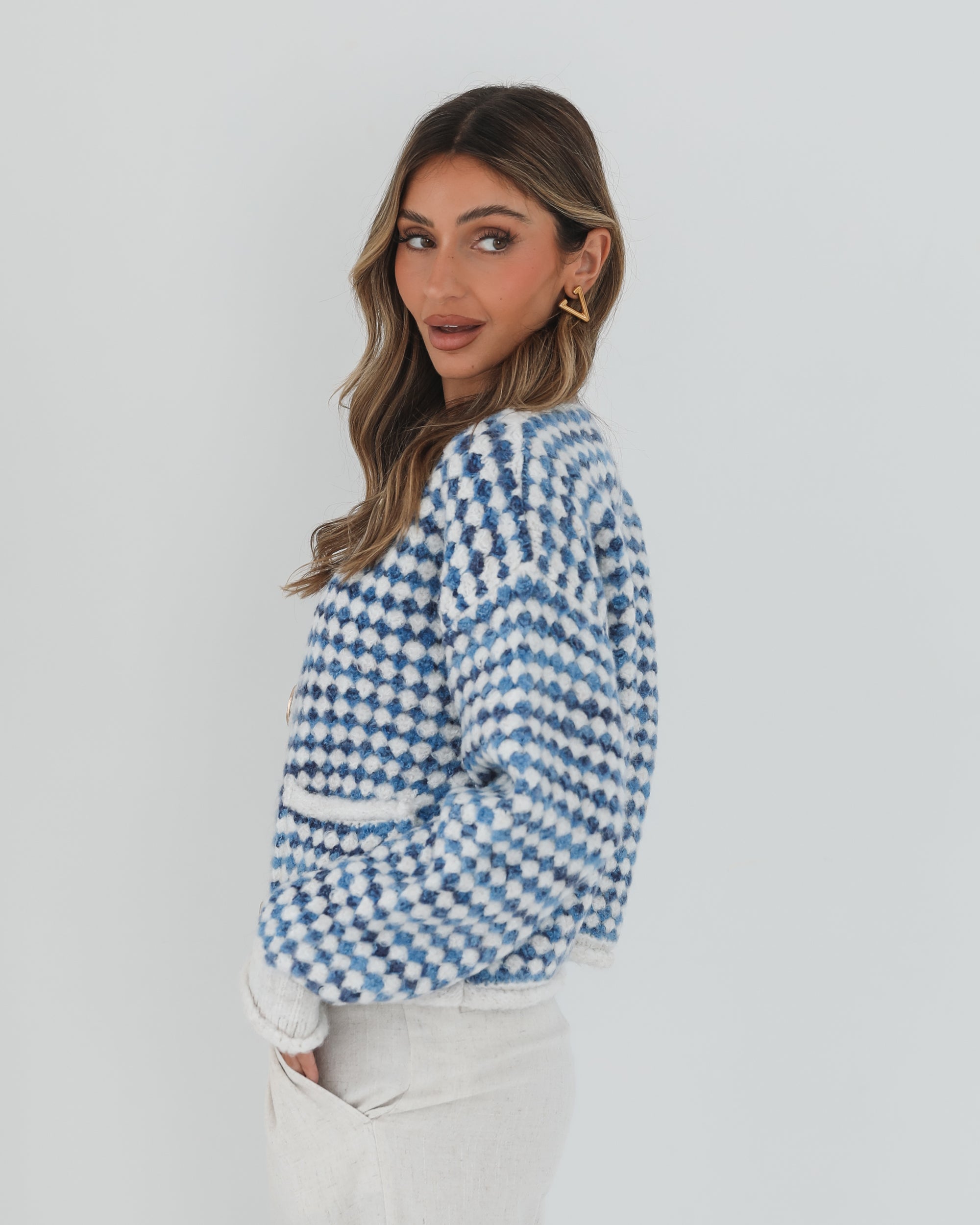 A woman with long, wavy brown hair stands against a plain white background, wearing a cozy blue and white checkered cardigan with oversized gold buttons and front pockets. She pairs it with light cream-colored pants and has her hands casually tucked into her pockets, giving a confident and composed look.