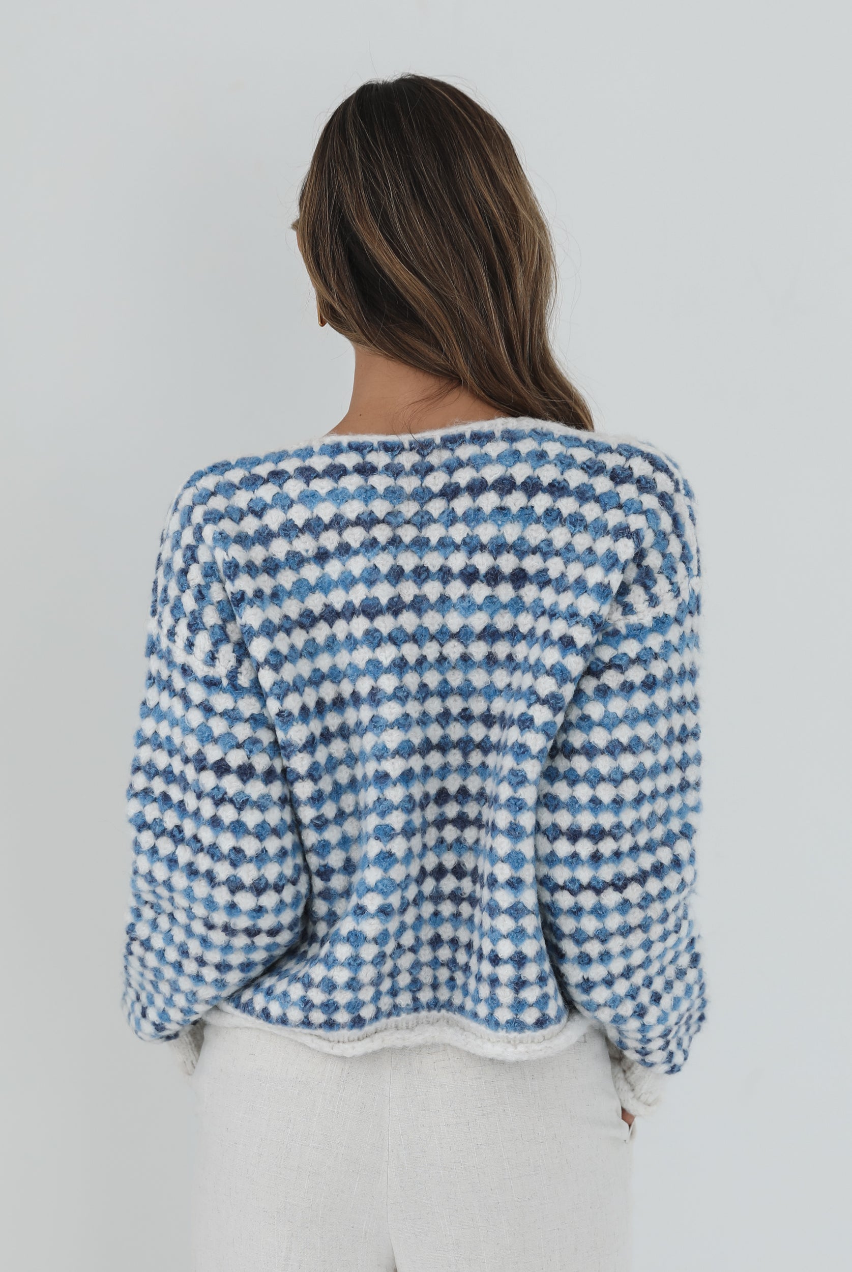 A woman with long, wavy brown hair stands against a plain white background, wearing a cozy blue and white checkered cardigan with oversized gold buttons and front pockets. She pairs it with light cream-colored pants and has her hands casually tucked into her pockets, giving a confident and composed look.