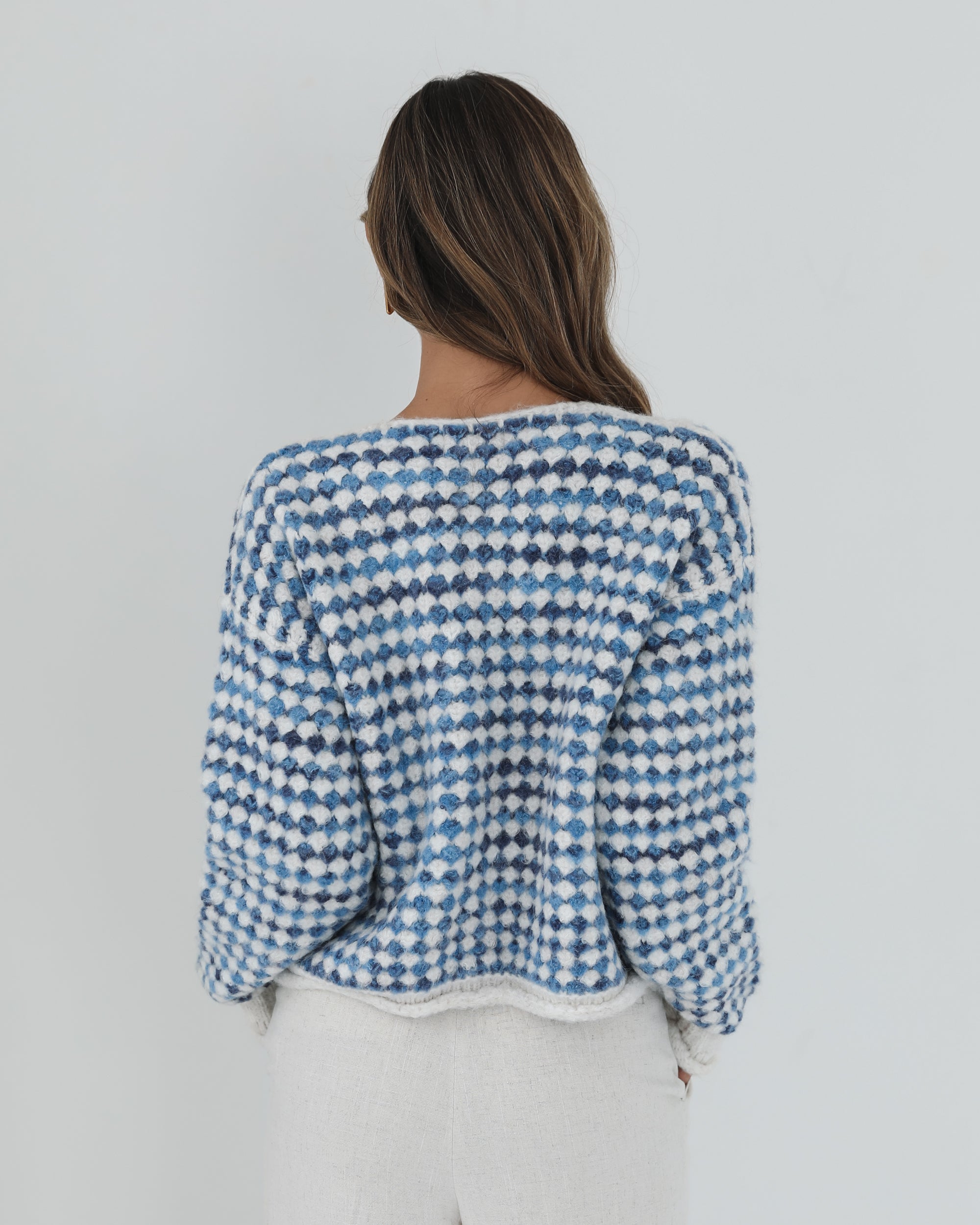A woman with long, wavy brown hair stands against a plain white background, wearing a cozy blue and white checkered cardigan with oversized gold buttons and front pockets. She pairs it with light cream-colored pants and has her hands casually tucked into her pockets, giving a confident and composed look.