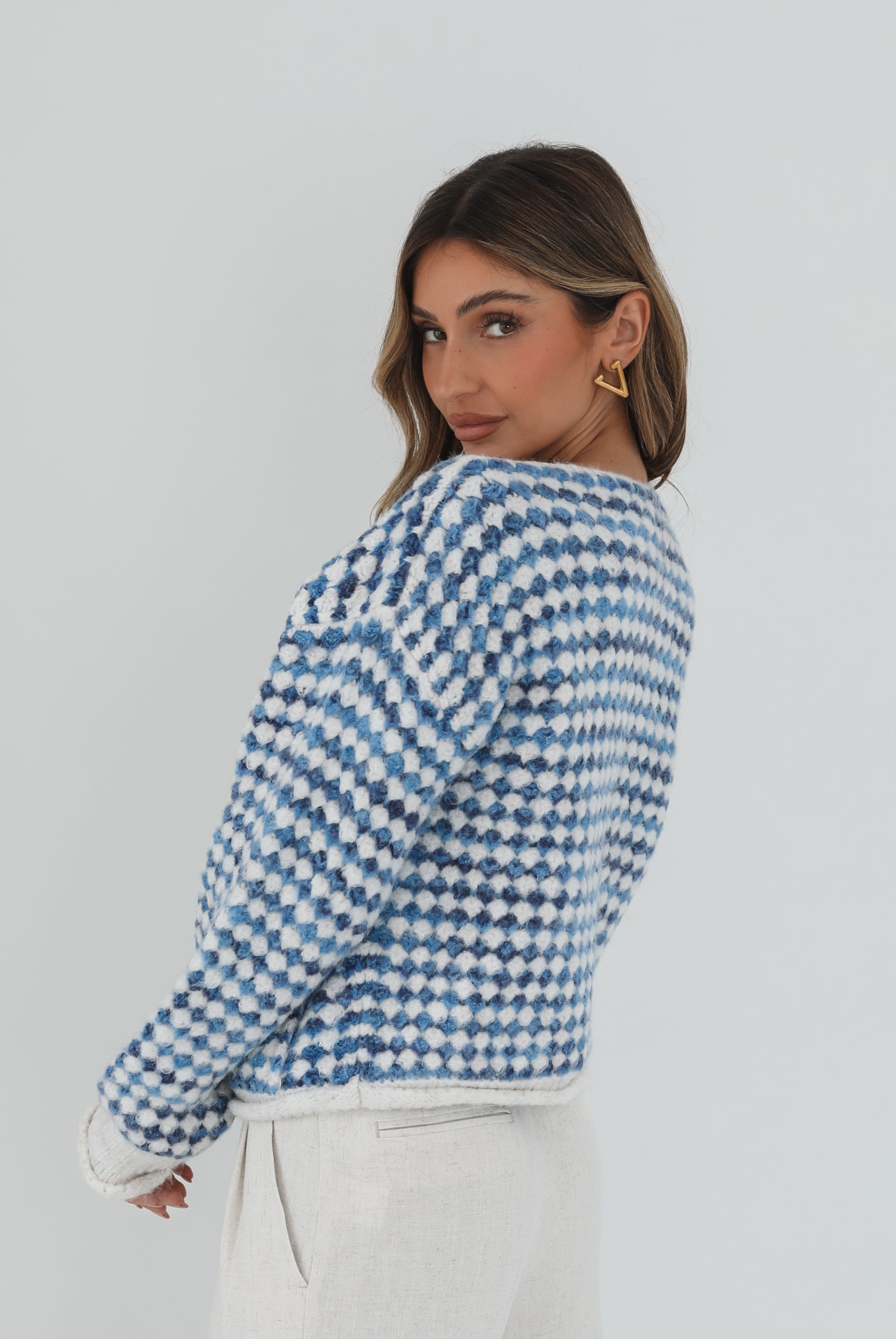 A woman with long, wavy brown hair stands against a plain white background, wearing a cozy blue and white checkered cardigan with oversized gold buttons and front pockets. She pairs it with light cream-colored pants and has her hands casually tucked into her pockets, giving a confident and composed look.