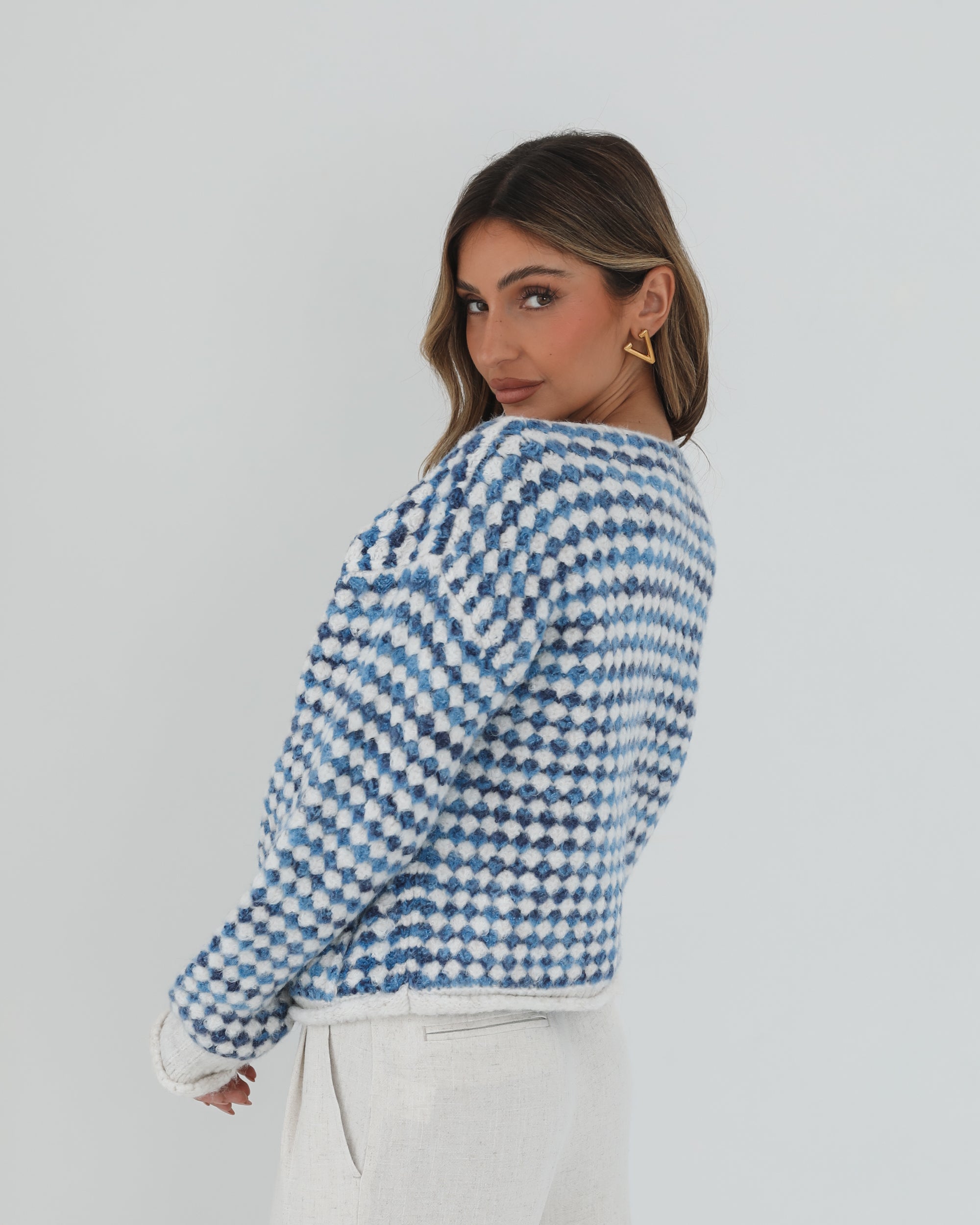 A woman with long, wavy brown hair stands against a plain white background, wearing a cozy blue and white checkered cardigan with oversized gold buttons and front pockets. She pairs it with light cream-colored pants and has her hands casually tucked into her pockets, giving a confident and composed look.