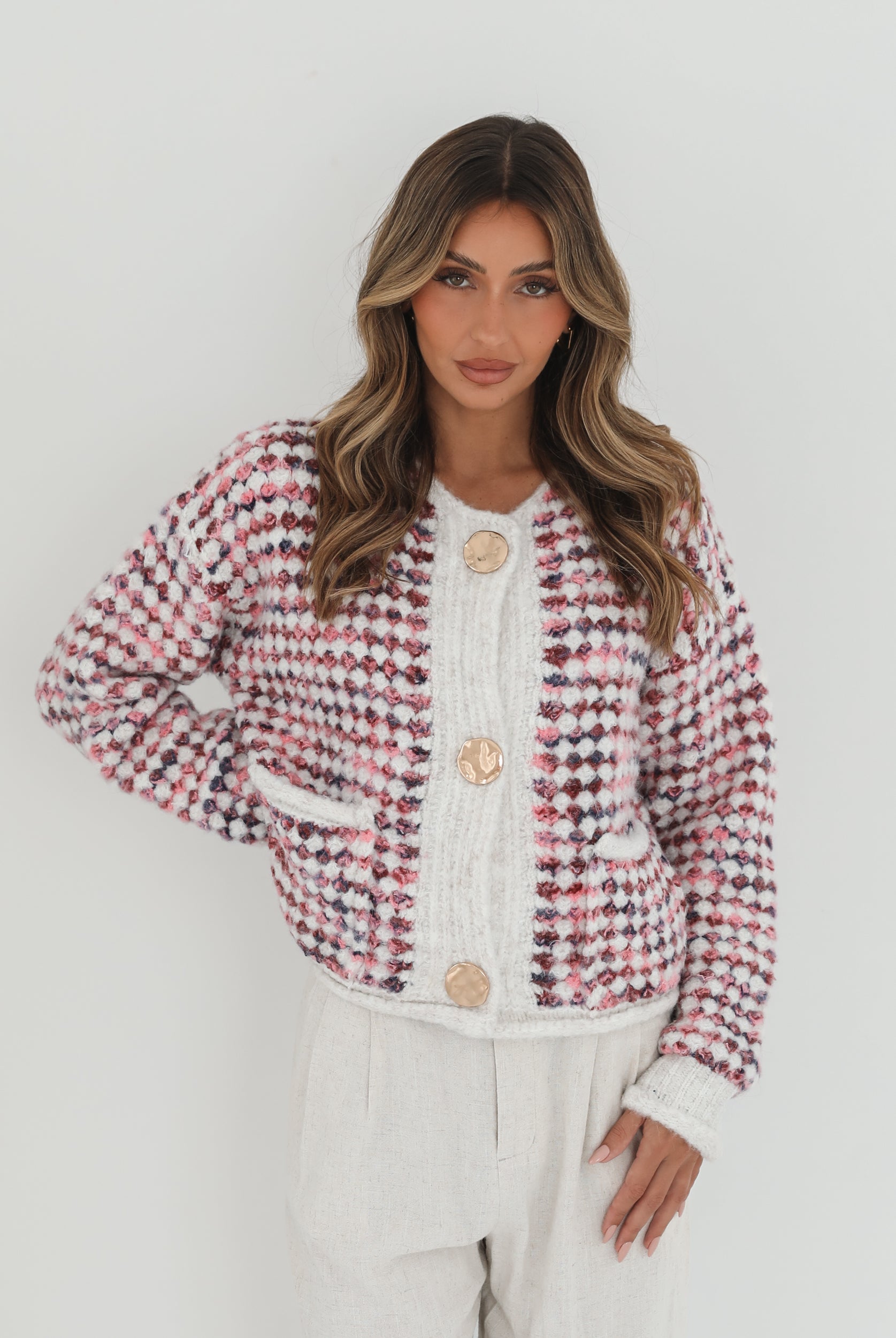 A woman with long, wavy brown hair stands against a plain white background, wearing a textured cardigan with a red, pink, and navy checkered pattern. The cardigan features large gold buttons and front pockets, layered over a white knit top. She pairs it with light cream-colored pants and poses with her hands in her pockets, gazing slightly to the side with a composed expression.