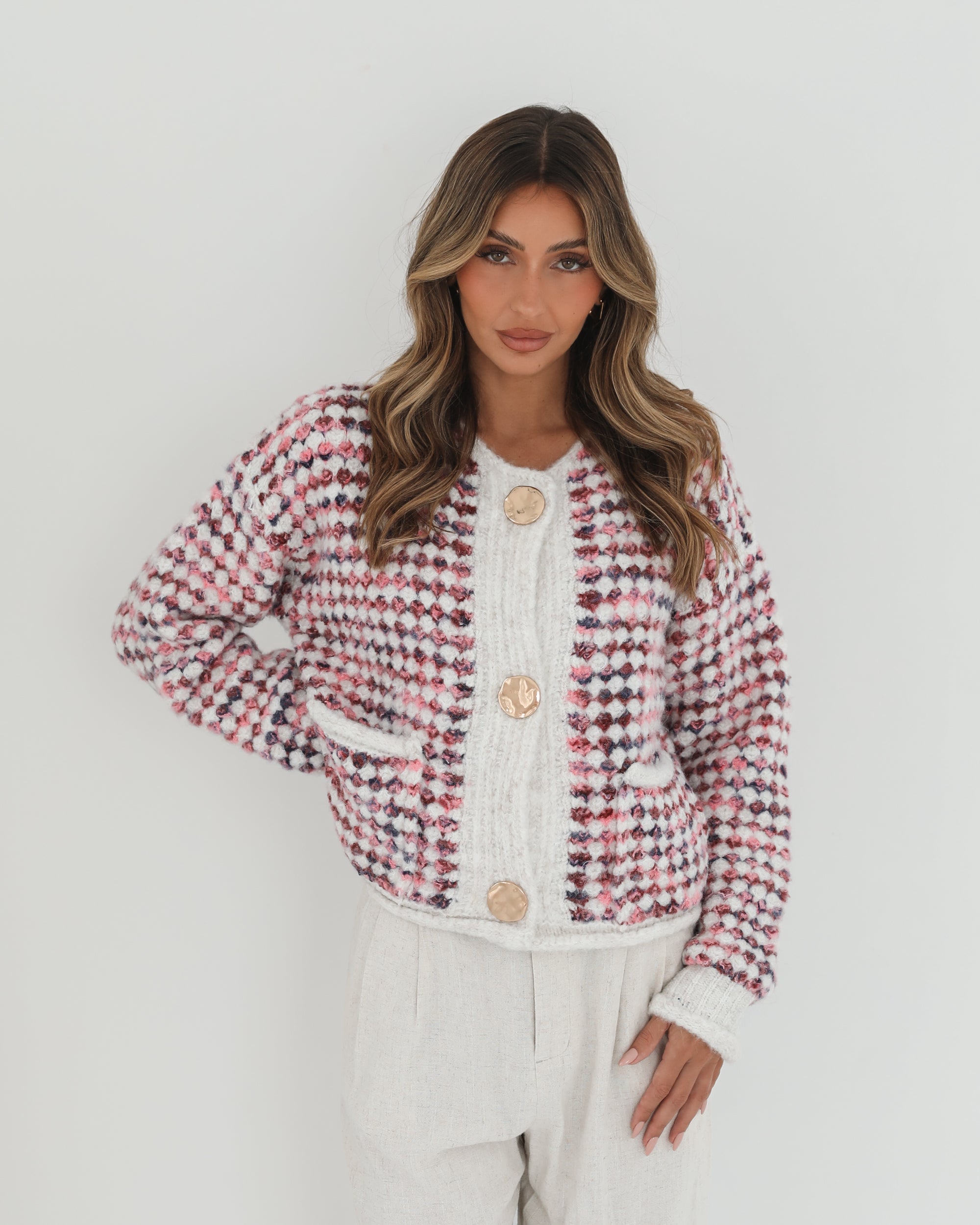 A woman with long, wavy brown hair stands against a plain white background, wearing a textured cardigan with a red, pink, and navy checkered pattern. The cardigan features large gold buttons and front pockets, layered over a white knit top. She pairs it with light cream-colored pants and poses with her hands in her pockets, gazing slightly to the side with a composed expression.