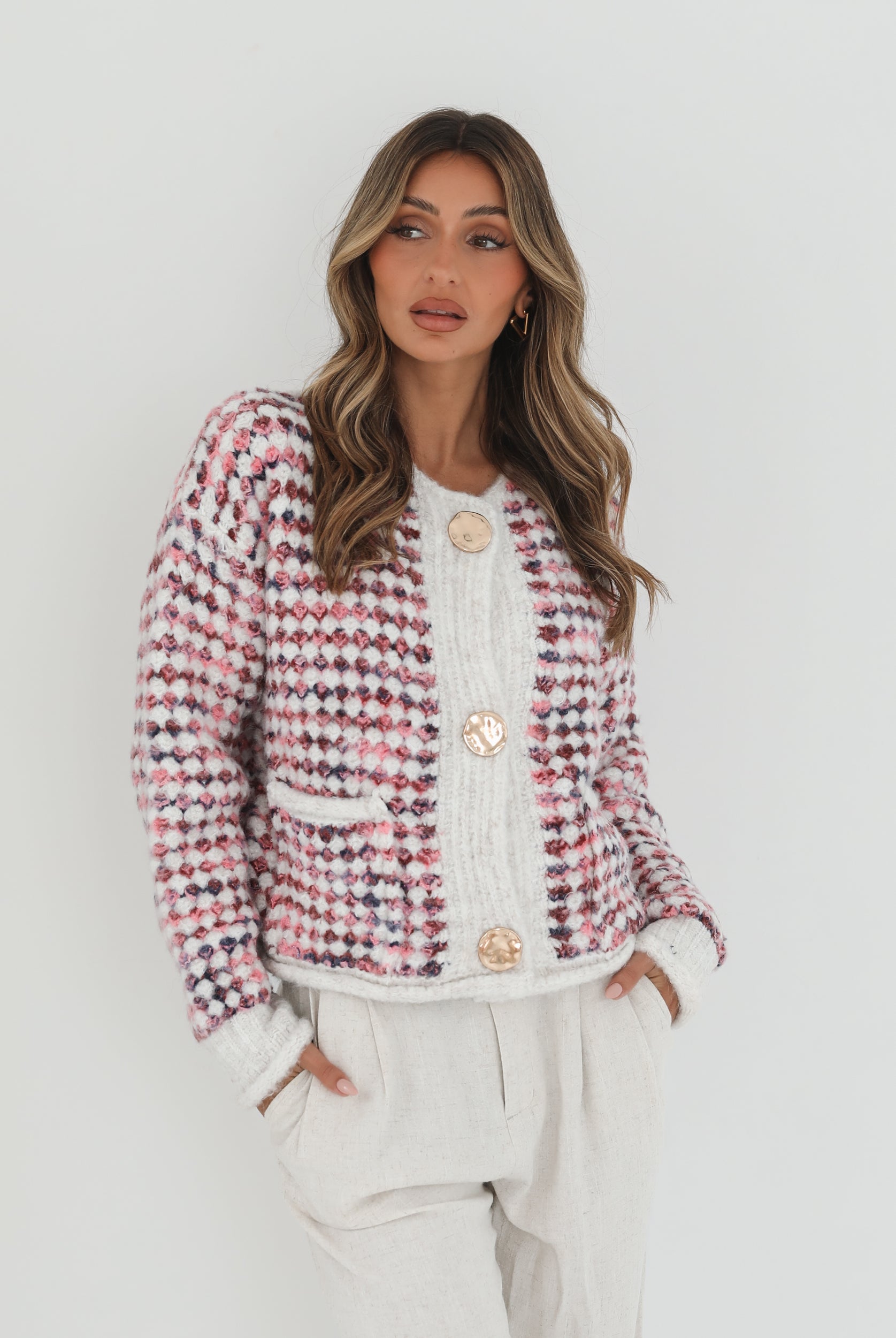 A woman with long, wavy brown hair stands against a plain white background, wearing a textured cardigan with a red, pink, and navy checkered pattern. The cardigan features large gold buttons and front pockets, layered over a white knit top. She pairs it with light cream-colored pants and poses with her hands in her pockets, gazing slightly to the side with a composed expression.