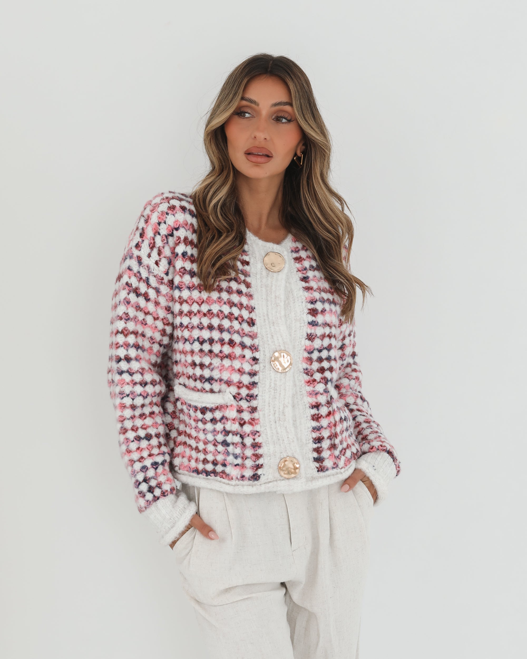 A woman with long, wavy brown hair stands against a plain white background, wearing a textured cardigan with a red, pink, and navy checkered pattern. The cardigan features large gold buttons and front pockets, layered over a white knit top. She pairs it with light cream-colored pants and poses with her hands in her pockets, gazing slightly to the side with a composed expression.
