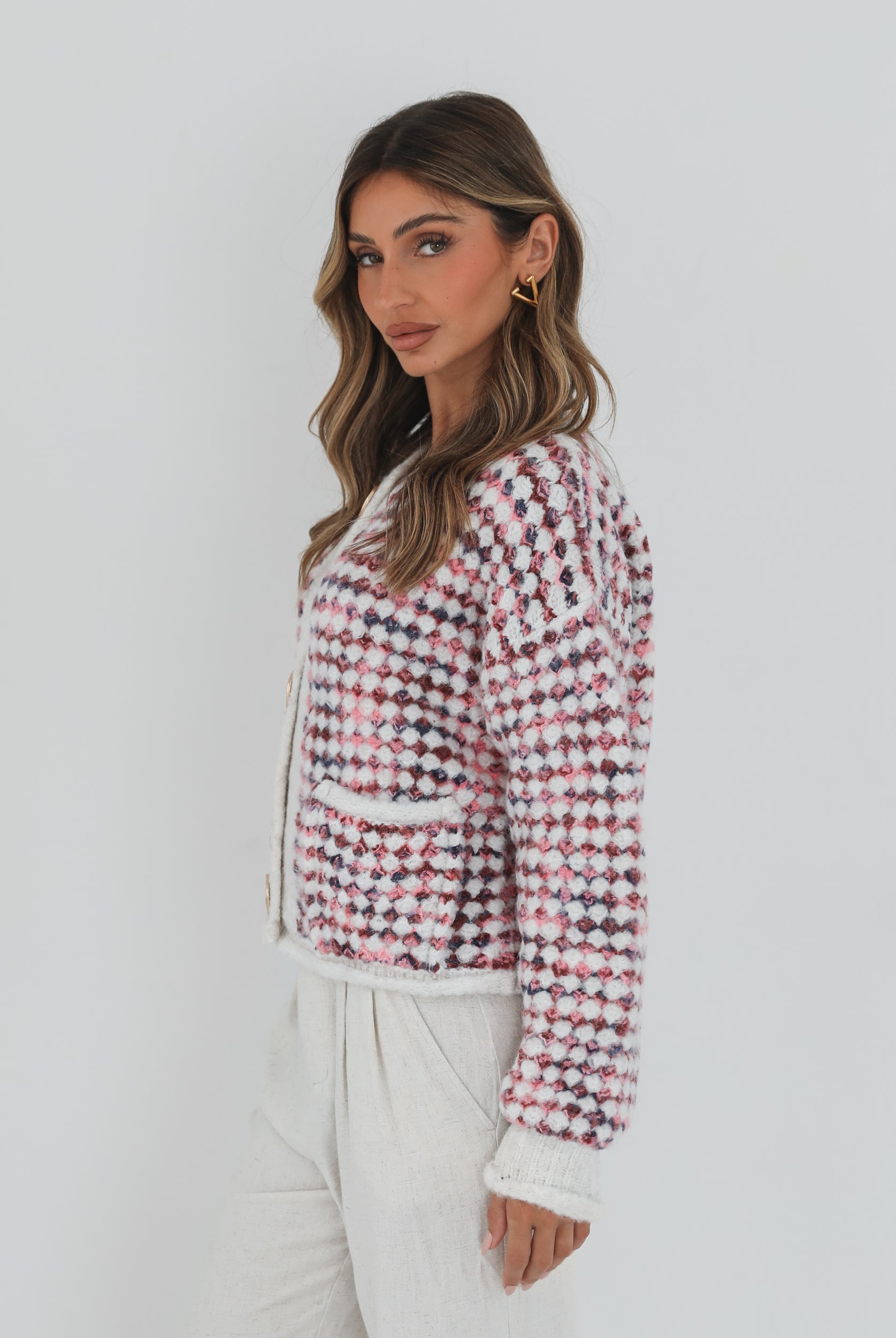 A woman with long, wavy brown hair stands against a plain white background, wearing a textured cardigan with a red, pink, and navy checkered pattern. The cardigan features large gold buttons and front pockets, layered over a white knit top. She pairs it with light cream-colored pants and poses with her hands in her pockets, gazing slightly to the side with a composed expression.