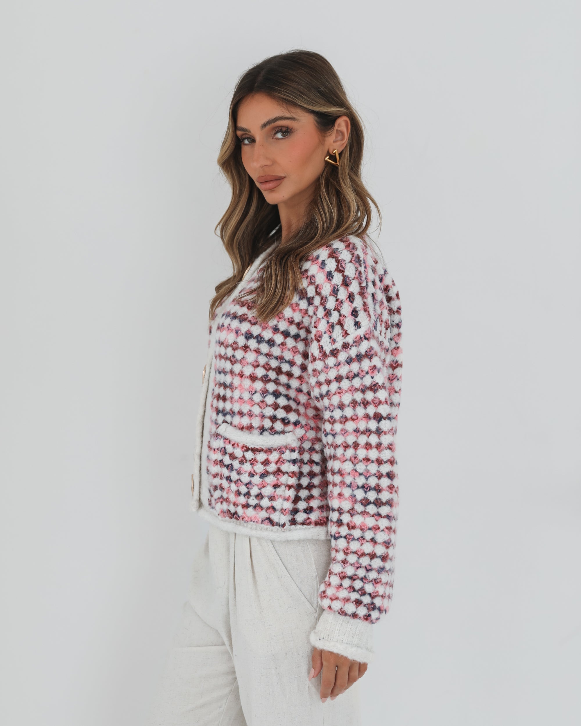 A woman with long, wavy brown hair stands against a plain white background, wearing a textured cardigan with a red, pink, and navy checkered pattern. The cardigan features large gold buttons and front pockets, layered over a white knit top. She pairs it with light cream-colored pants and poses with her hands in her pockets, gazing slightly to the side with a composed expression.