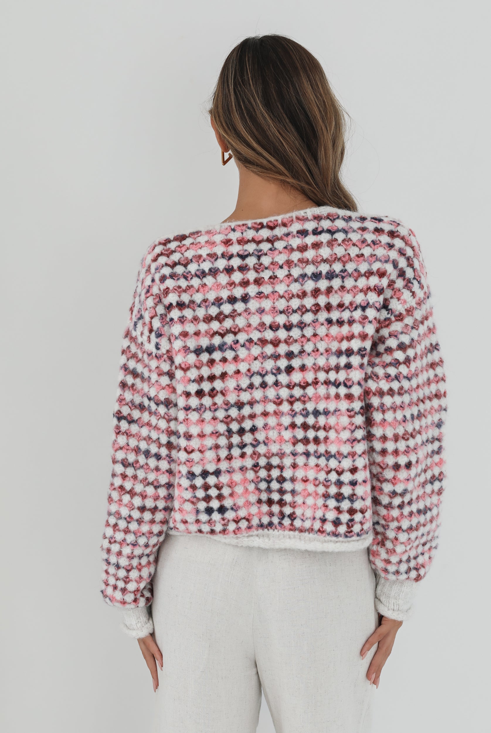 A woman with long, wavy brown hair stands against a plain white background, wearing a textured cardigan with a red, pink, and navy checkered pattern. The cardigan features large gold buttons and front pockets, layered over a white knit top. She pairs it with light cream-colored pants and poses with her hands in her pockets, gazing slightly to the side with a composed expression.