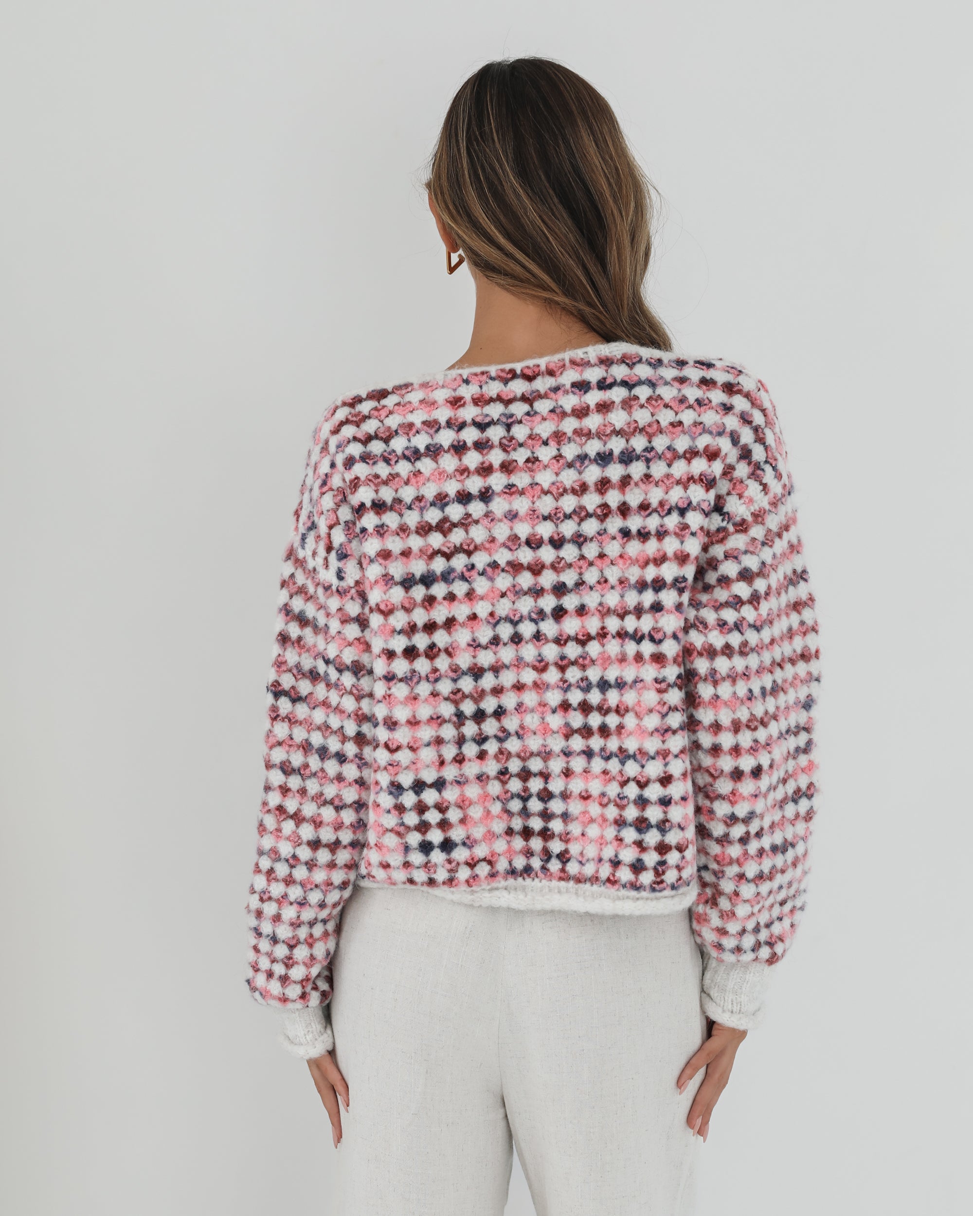 A woman with long, wavy brown hair stands against a plain white background, wearing a textured cardigan with a red, pink, and navy checkered pattern. The cardigan features large gold buttons and front pockets, layered over a white knit top. She pairs it with light cream-colored pants and poses with her hands in her pockets, gazing slightly to the side with a composed expression.