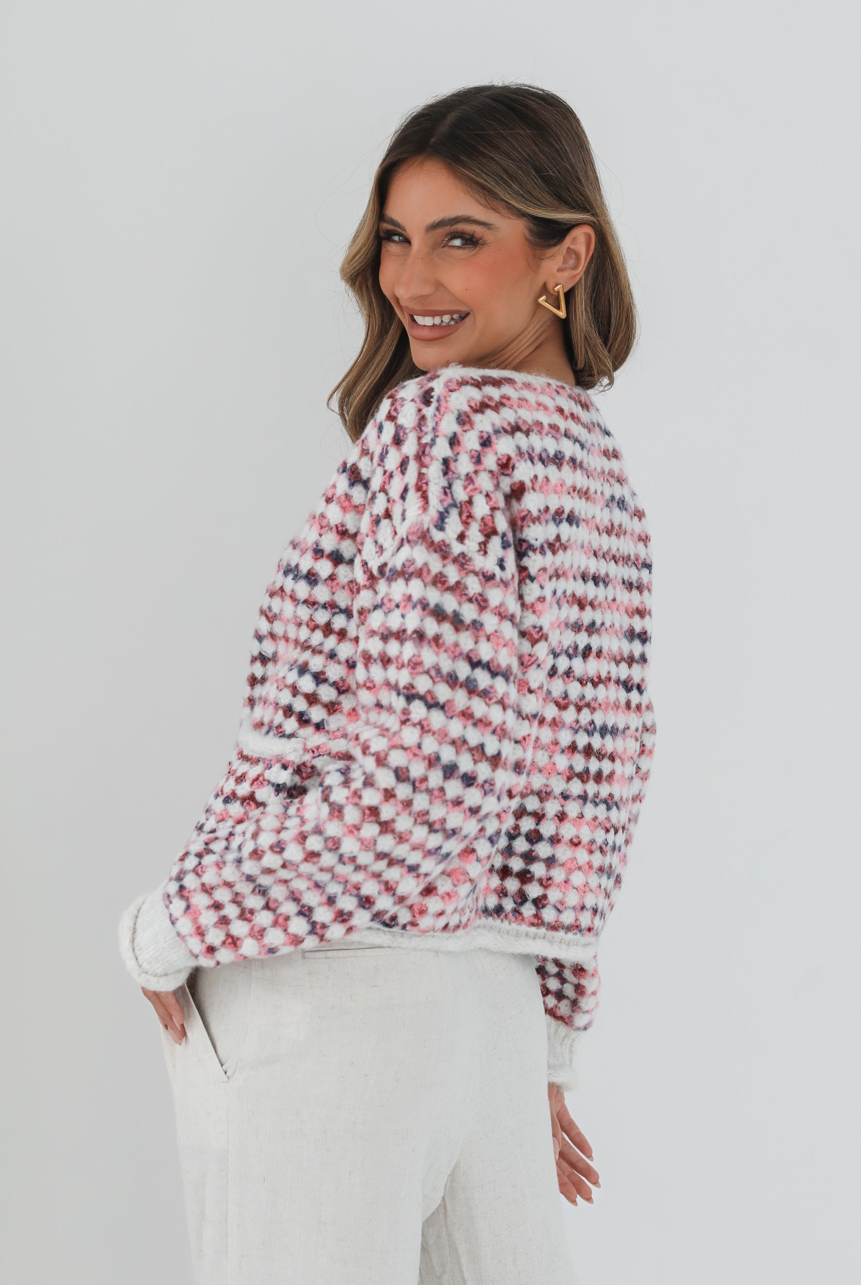 A woman with long, wavy brown hair stands against a plain white background, wearing a textured cardigan with a red, pink, and navy checkered pattern. The cardigan features large gold buttons and front pockets, layered over a white knit top. She pairs it with light cream-colored pants and poses with her hands in her pockets, gazing slightly to the side with a composed expression.