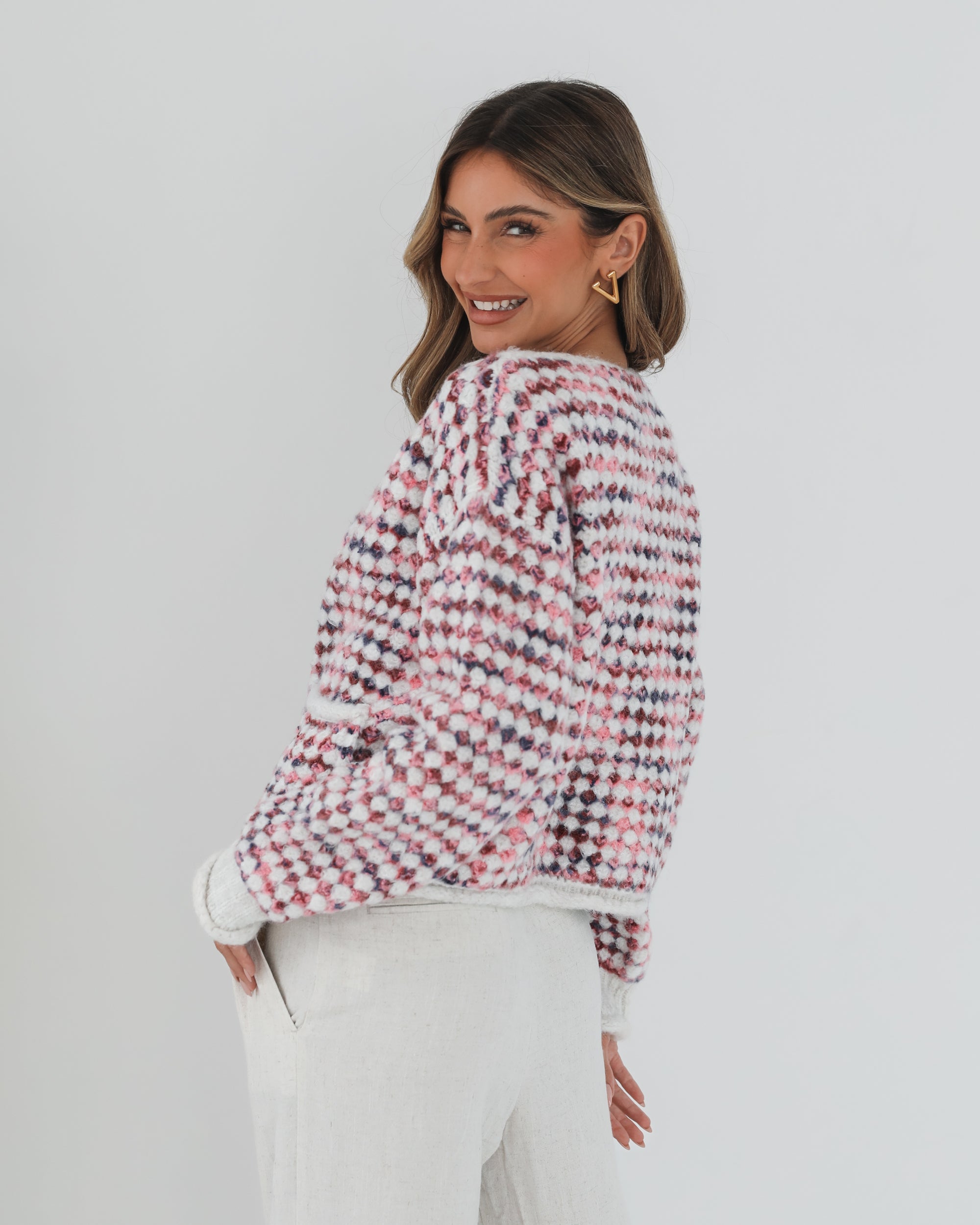 A woman with long, wavy brown hair stands against a plain white background, wearing a textured cardigan with a red, pink, and navy checkered pattern. The cardigan features large gold buttons and front pockets, layered over a white knit top. She pairs it with light cream-colored pants and poses with her hands in her pockets, gazing slightly to the side with a composed expression.