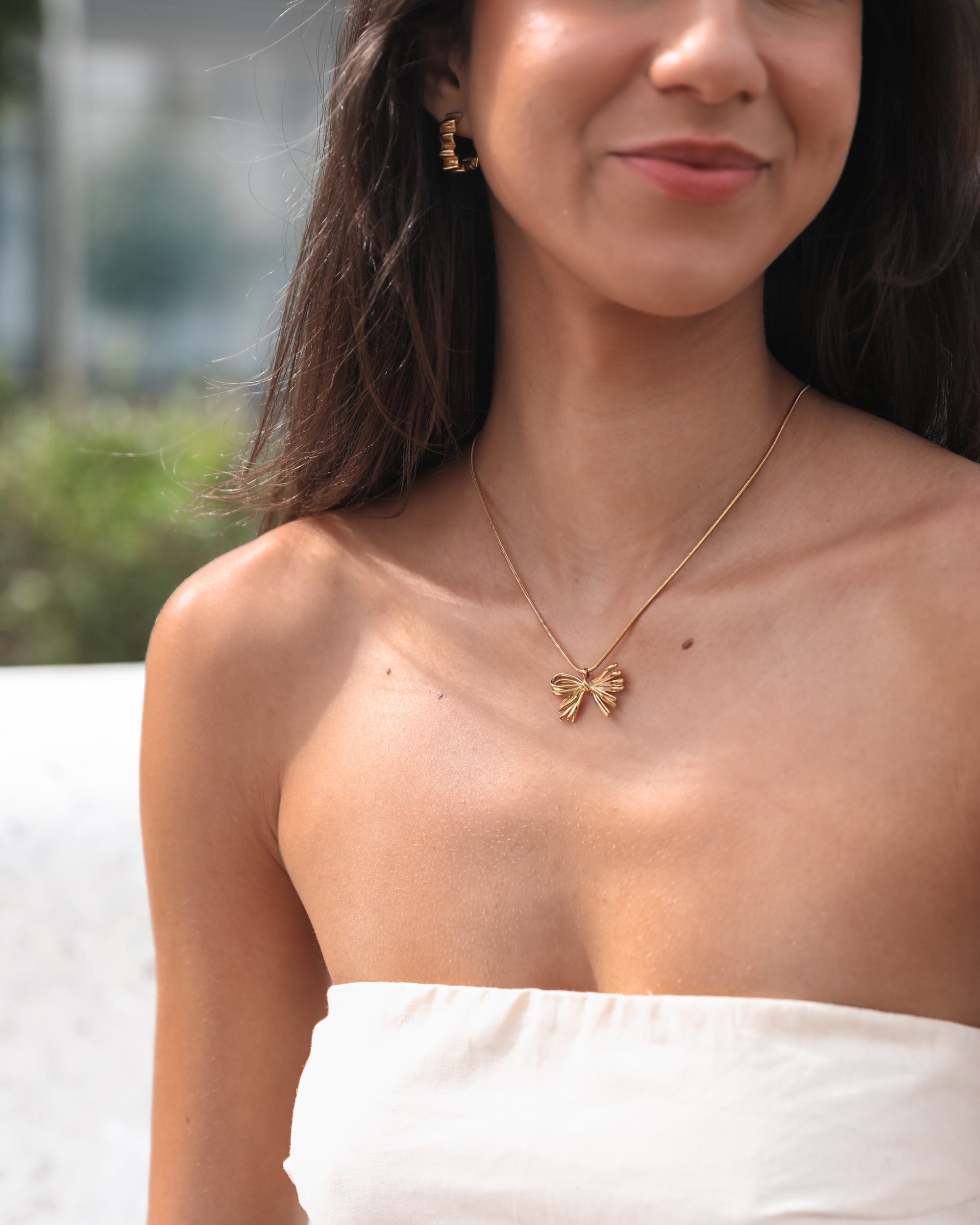 A close-up of a woman’s upper chest and neck, showcasing a delicate gold necklace with a bow-shaped pendant. She is wearing a white strapless top and small gold hoop earrings. The background is softly blurred, highlighting her smooth skin and subtle smile.