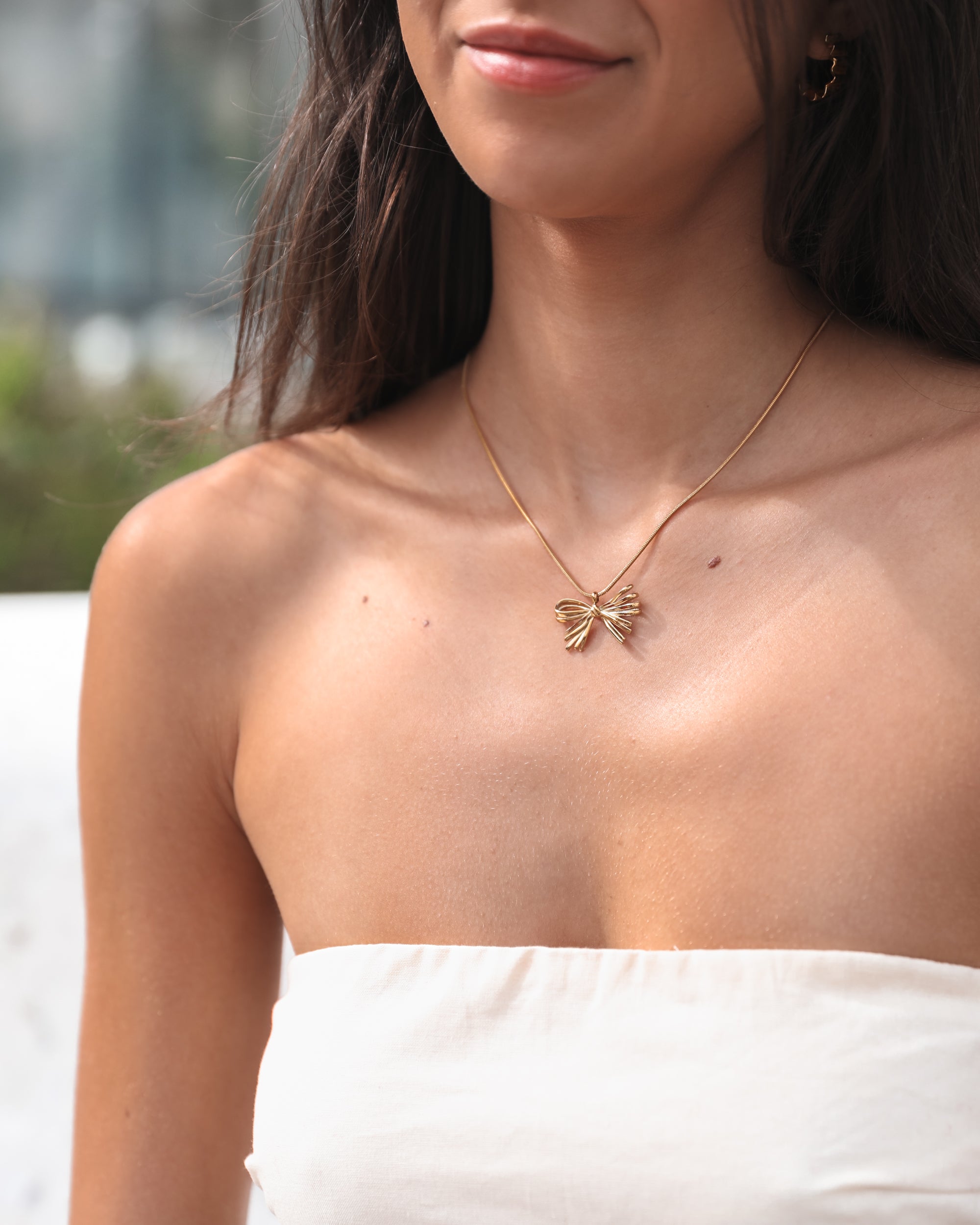 A close-up of a woman’s upper chest and neck, showcasing a delicate gold necklace with a bow-shaped pendant. She is wearing a white strapless top and small gold hoop earrings. The background is softly blurred, highlighting her smooth skin and subtle smile.