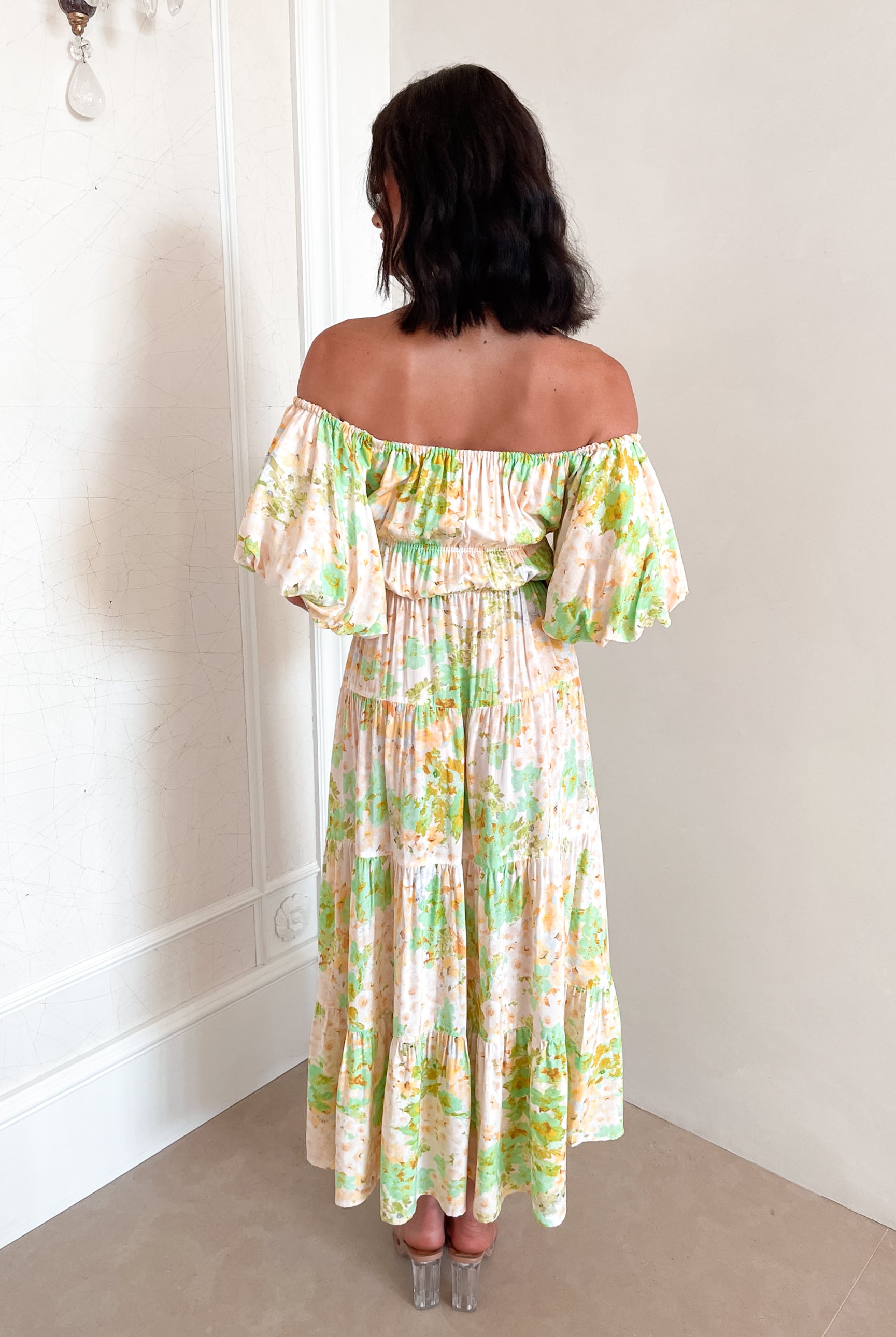 A woman with shoulder-length dark hair stands against a light beige wall, wearing an off-the-shoulder, flowy midi dress with a pastel floral pattern in shades of yellow, green, and peach. The dress features puffed sleeves and a smocked waist. She completes the look with clear strap heels and a delicate necklace, posing gracefully with her hands gently clasped in front.