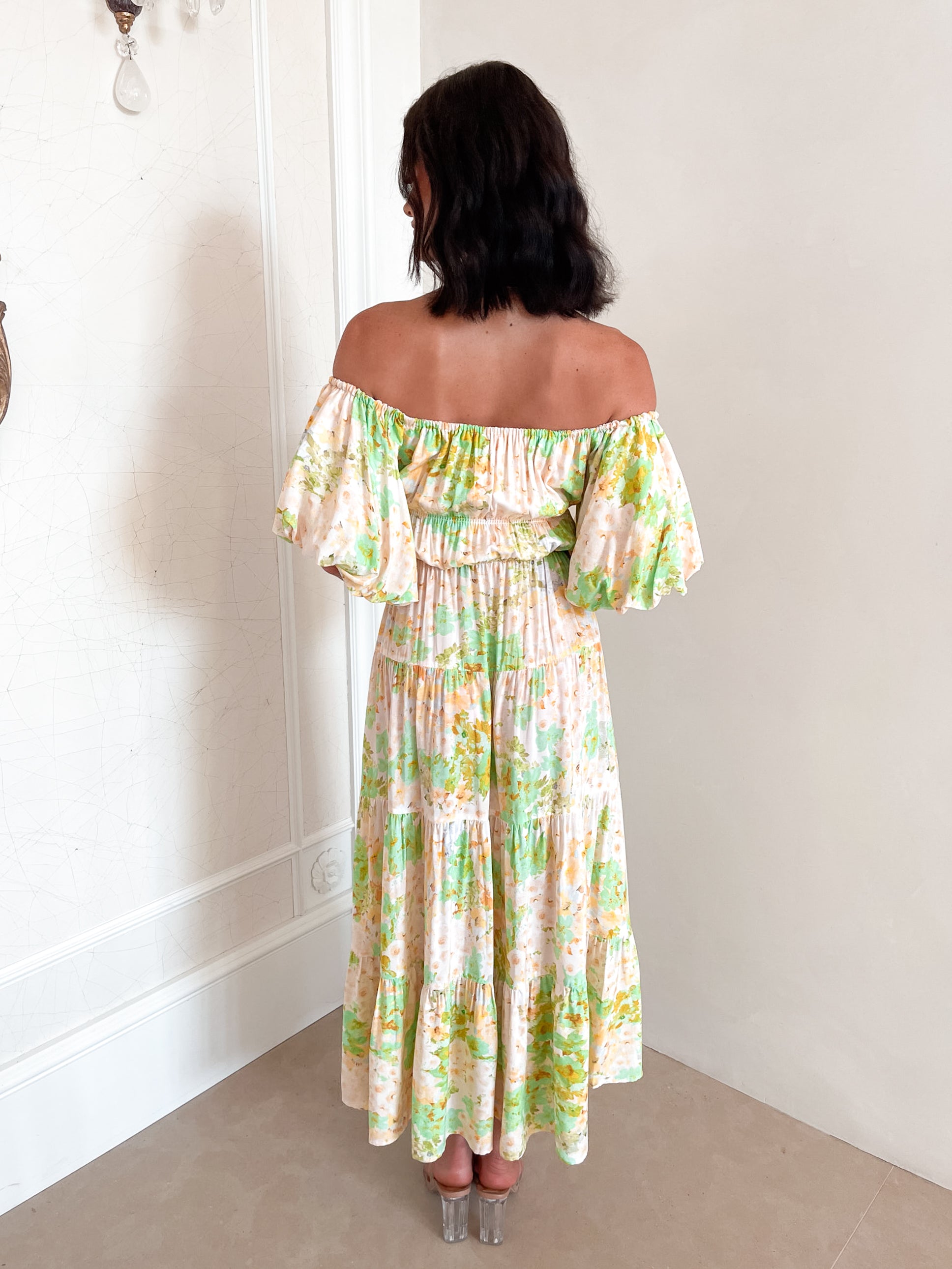 A woman with shoulder-length dark hair stands against a light beige wall, wearing an off-the-shoulder, flowy midi dress with a pastel floral pattern in shades of yellow, green, and peach. The dress features puffed sleeves and a smocked waist. She completes the look with clear strap heels and a delicate necklace, posing gracefully with her hands gently clasped in front.