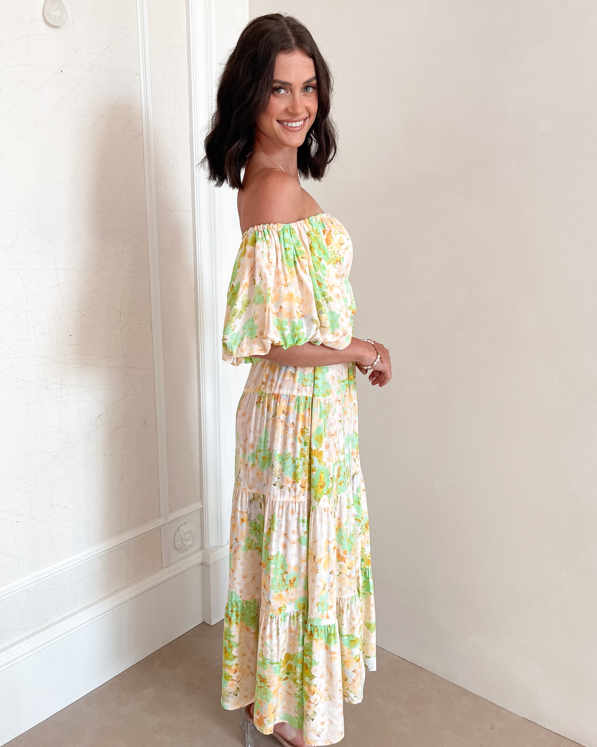 A woman with shoulder-length dark hair stands against a light beige wall, wearing an off-the-shoulder, flowy midi dress with a pastel floral pattern in shades of yellow, green, and peach. The dress features puffed sleeves and a smocked waist. She completes the look with clear strap heels and a delicate necklace, posing gracefully with her hands gently clasped in front.