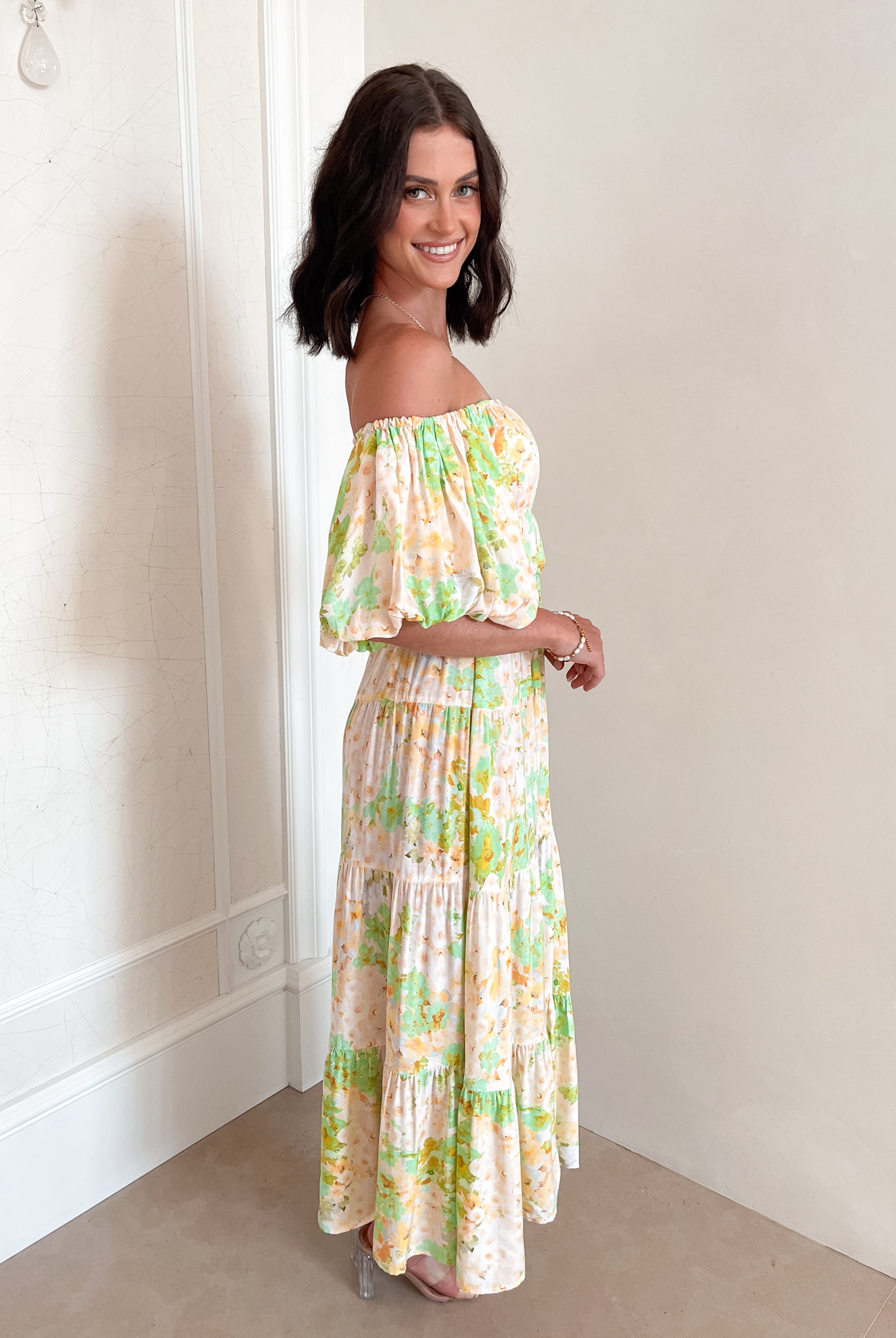 A woman with shoulder-length dark hair stands against a light beige wall, wearing an off-the-shoulder, flowy midi dress with a pastel floral pattern in shades of yellow, green, and peach. The dress features puffed sleeves and a smocked waist. She completes the look with clear strap heels and a delicate necklace, posing gracefully with her hands gently clasped in front.