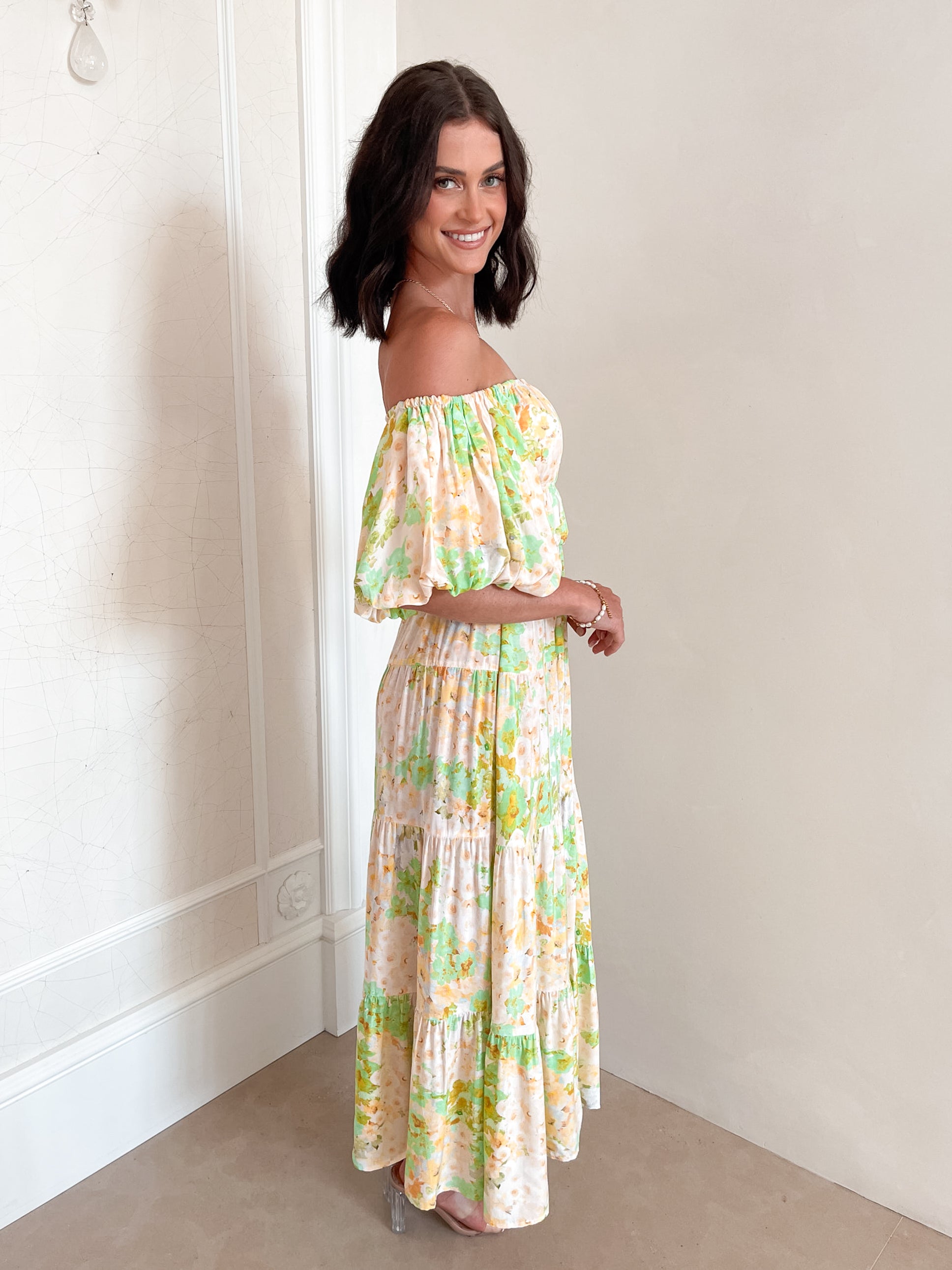 A woman with shoulder-length dark hair stands against a light beige wall, wearing an off-the-shoulder, flowy midi dress with a pastel floral pattern in shades of yellow, green, and peach. The dress features puffed sleeves and a smocked waist. She completes the look with clear strap heels and a delicate necklace, posing gracefully with her hands gently clasped in front.