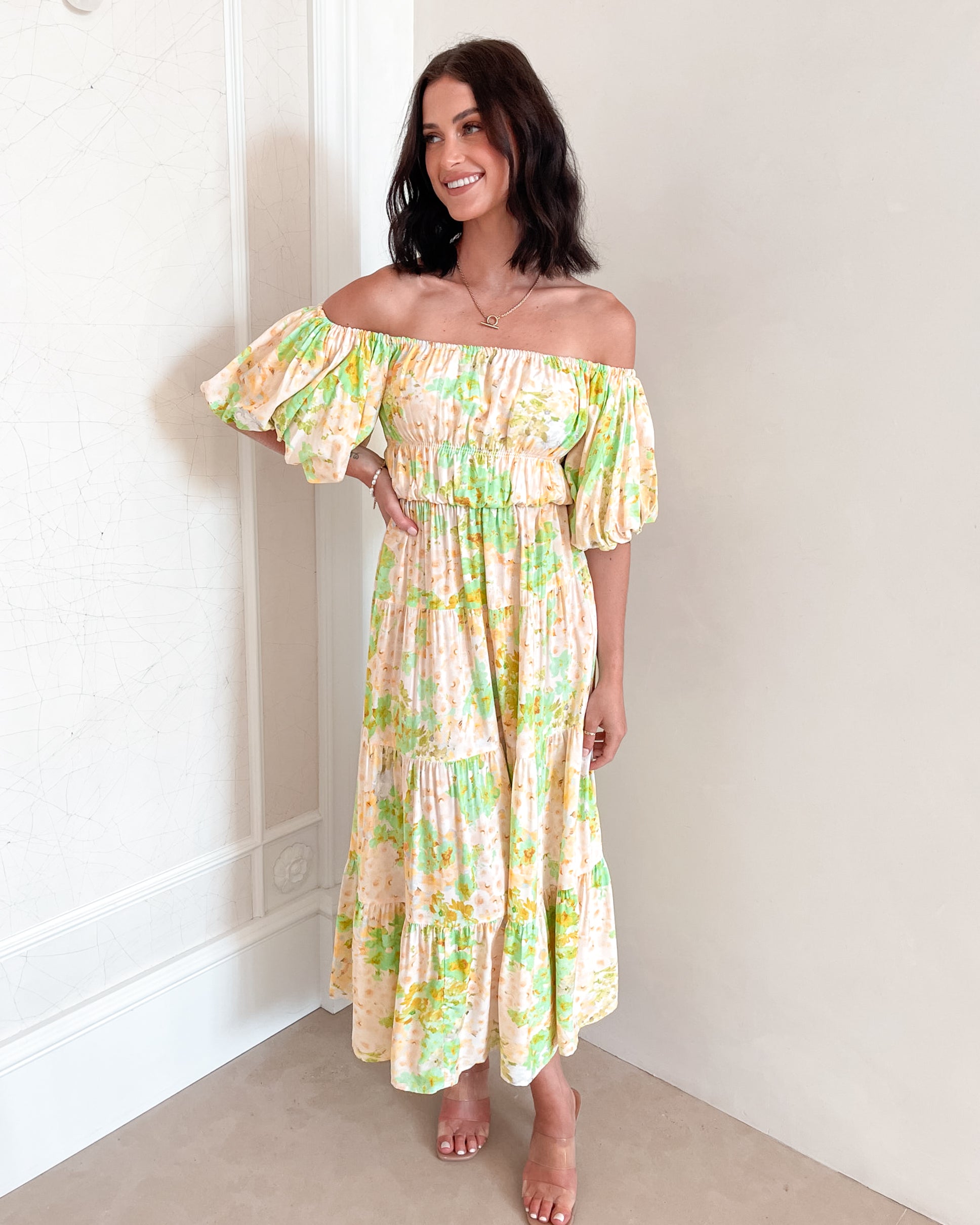 A woman with shoulder-length dark hair stands against a light beige wall, wearing an off-the-shoulder, flowy midi dress with a pastel floral pattern in shades of yellow, green, and peach. The dress features puffed sleeves and a smocked waist. She completes the look with clear strap heels and a delicate necklace, posing gracefully with her hands gently clasped in front.