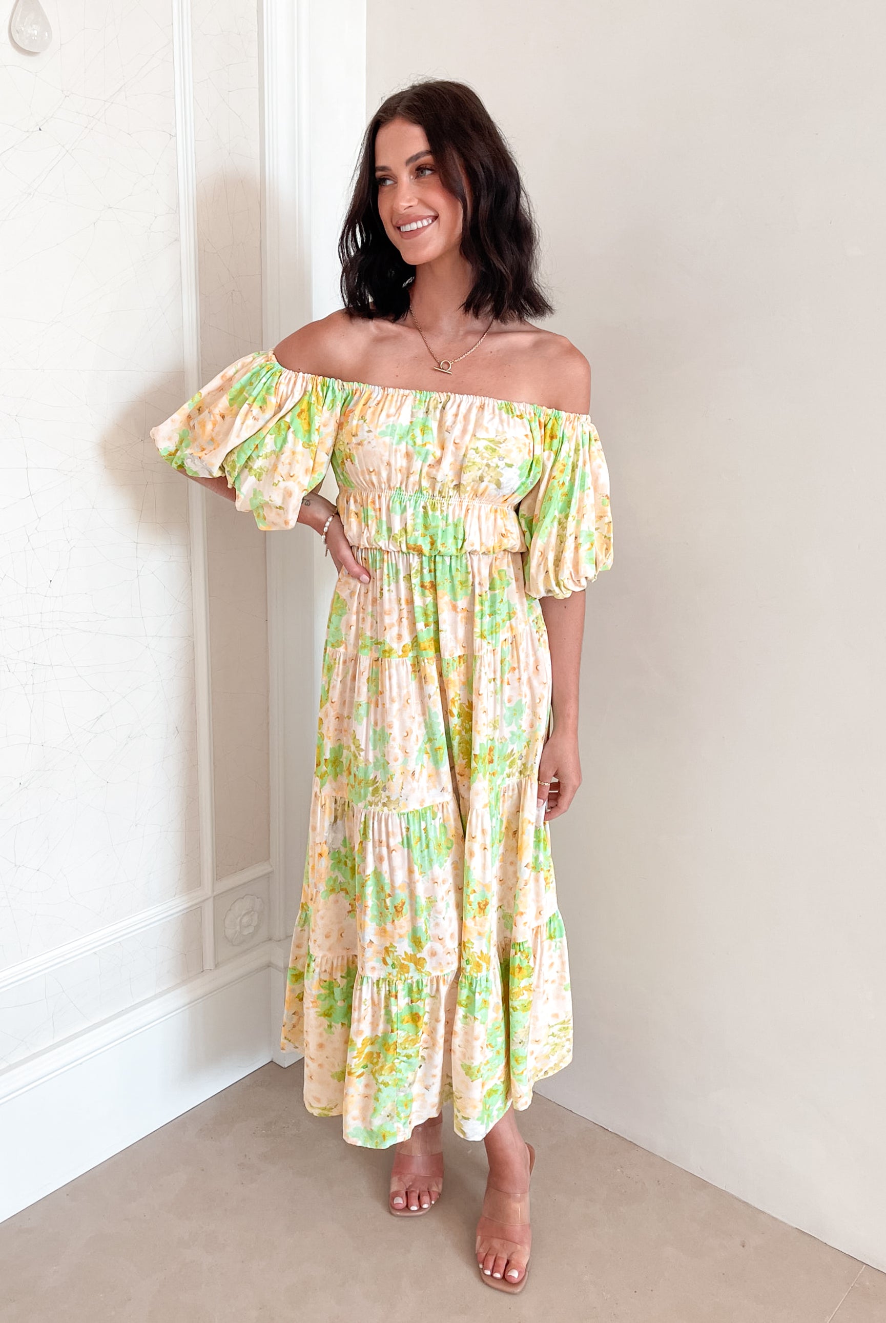 A woman with shoulder-length dark hair stands against a light beige wall, wearing an off-the-shoulder, flowy midi dress with a pastel floral pattern in shades of yellow, green, and peach. The dress features puffed sleeves and a smocked waist. She completes the look with clear strap heels and a delicate necklace, posing gracefully with her hands gently clasped in front.