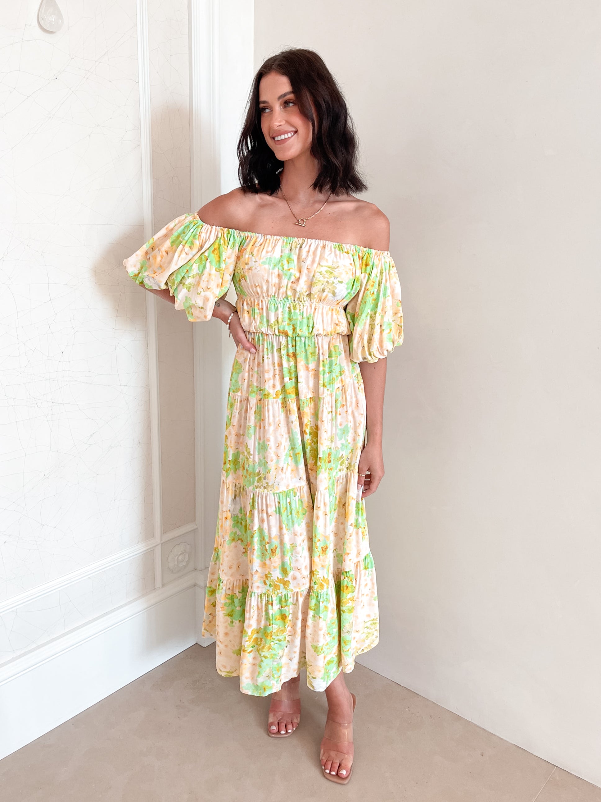 A woman with shoulder-length dark hair stands against a light beige wall, wearing an off-the-shoulder, flowy midi dress with a pastel floral pattern in shades of yellow, green, and peach. The dress features puffed sleeves and a smocked waist. She completes the look with clear strap heels and a delicate necklace, posing gracefully with her hands gently clasped in front.