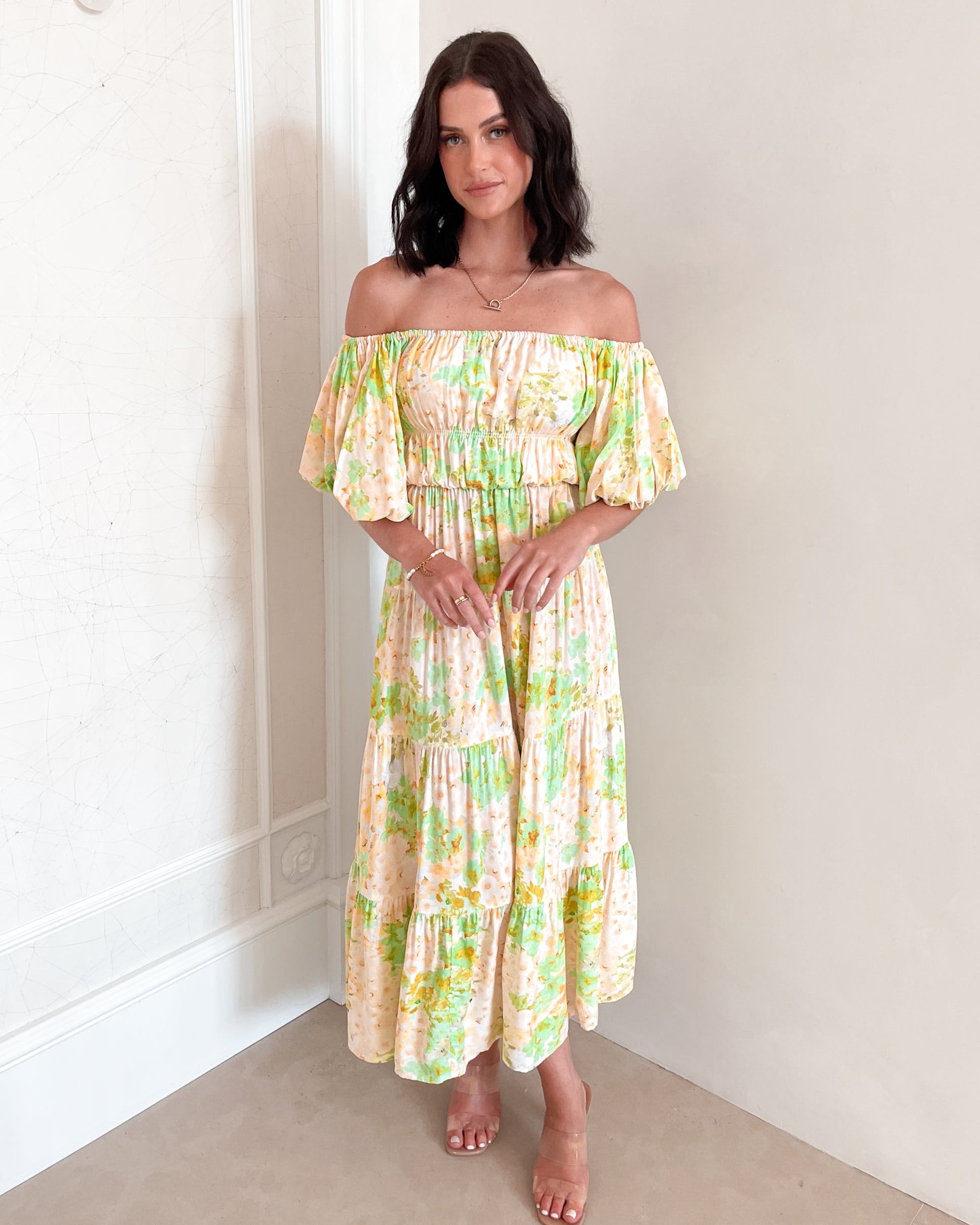 A woman with shoulder-length dark hair stands against a light beige wall, wearing an off-the-shoulder, flowy midi dress with a pastel floral pattern in shades of yellow, green, and peach. The dress features puffed sleeves and a smocked waist. She completes the look with clear strap heels and a delicate necklace, posing gracefully with her hands gently clasped in front.