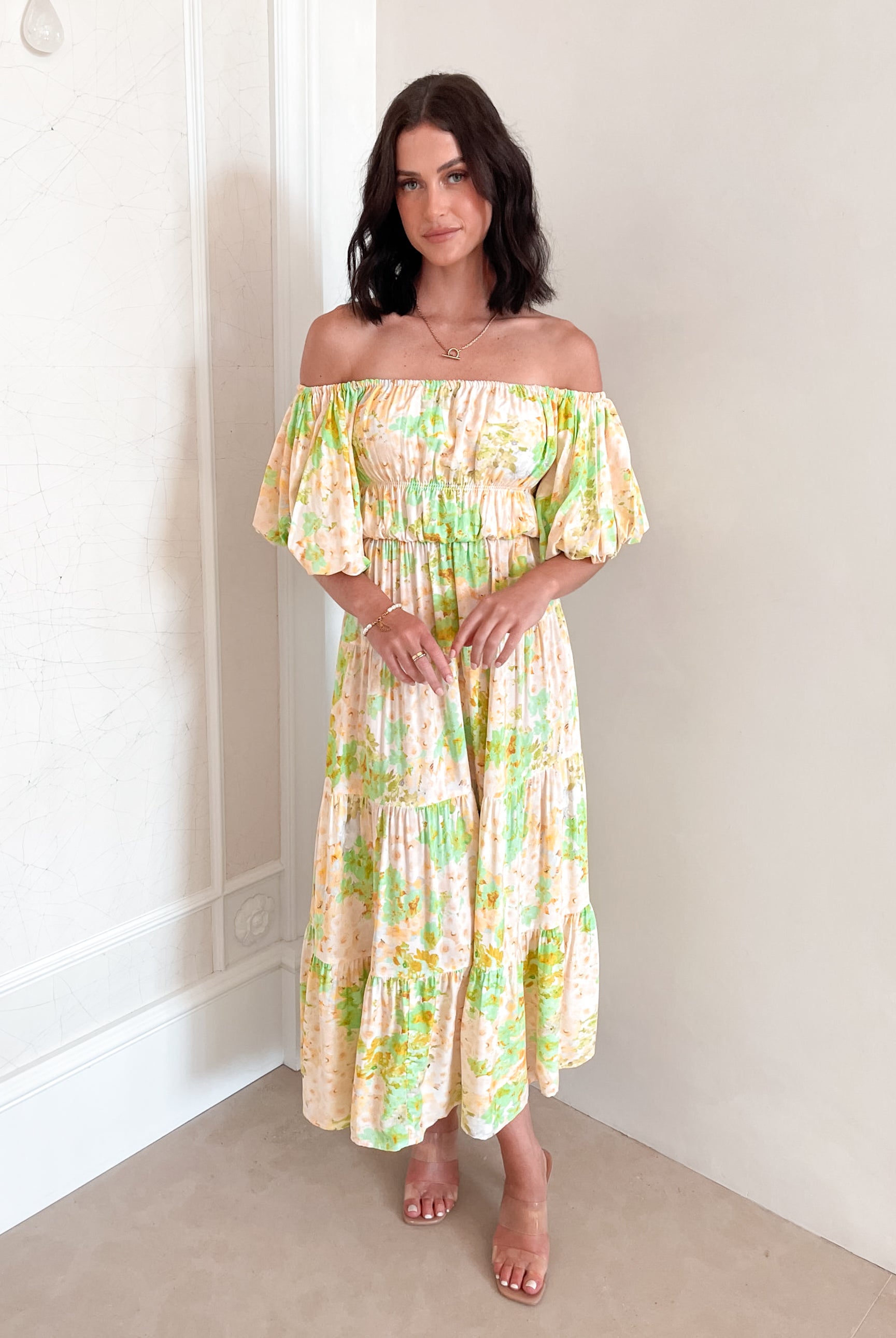 A woman with shoulder-length dark hair stands against a light beige wall, wearing an off-the-shoulder, flowy midi dress with a pastel floral pattern in shades of yellow, green, and peach. The dress features puffed sleeves and a smocked waist. She completes the look with clear strap heels and a delicate necklace, posing gracefully with her hands gently clasped in front.