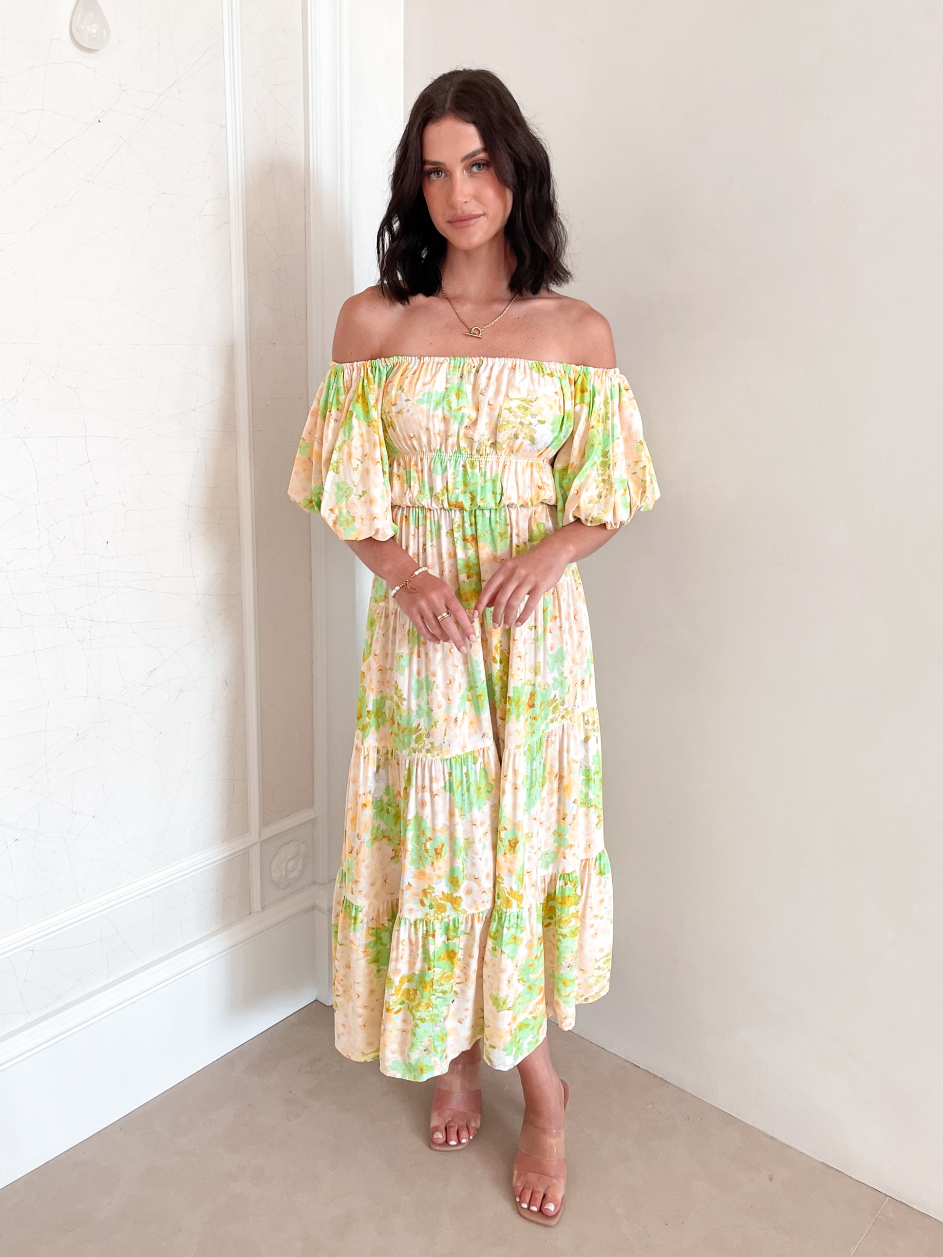 A woman with shoulder-length dark hair stands against a light beige wall, wearing an off-the-shoulder, flowy midi dress with a pastel floral pattern in shades of yellow, green, and peach. The dress features puffed sleeves and a smocked waist. She completes the look with clear strap heels and a delicate necklace, posing gracefully with her hands gently clasped in front.