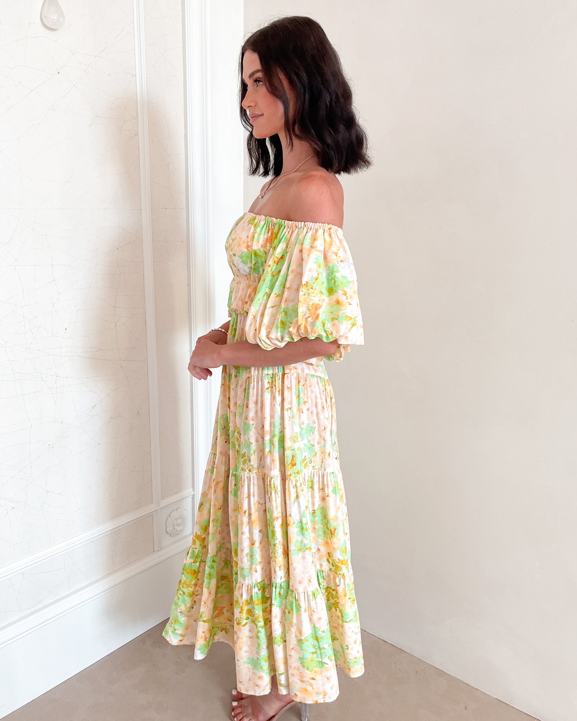 A woman with shoulder-length dark hair stands against a light beige wall, wearing an off-the-shoulder, flowy midi dress with a pastel floral pattern in shades of yellow, green, and peach. The dress features puffed sleeves and a smocked waist. She completes the look with clear strap heels and a delicate necklace, posing gracefully with her hands gently clasped in front.