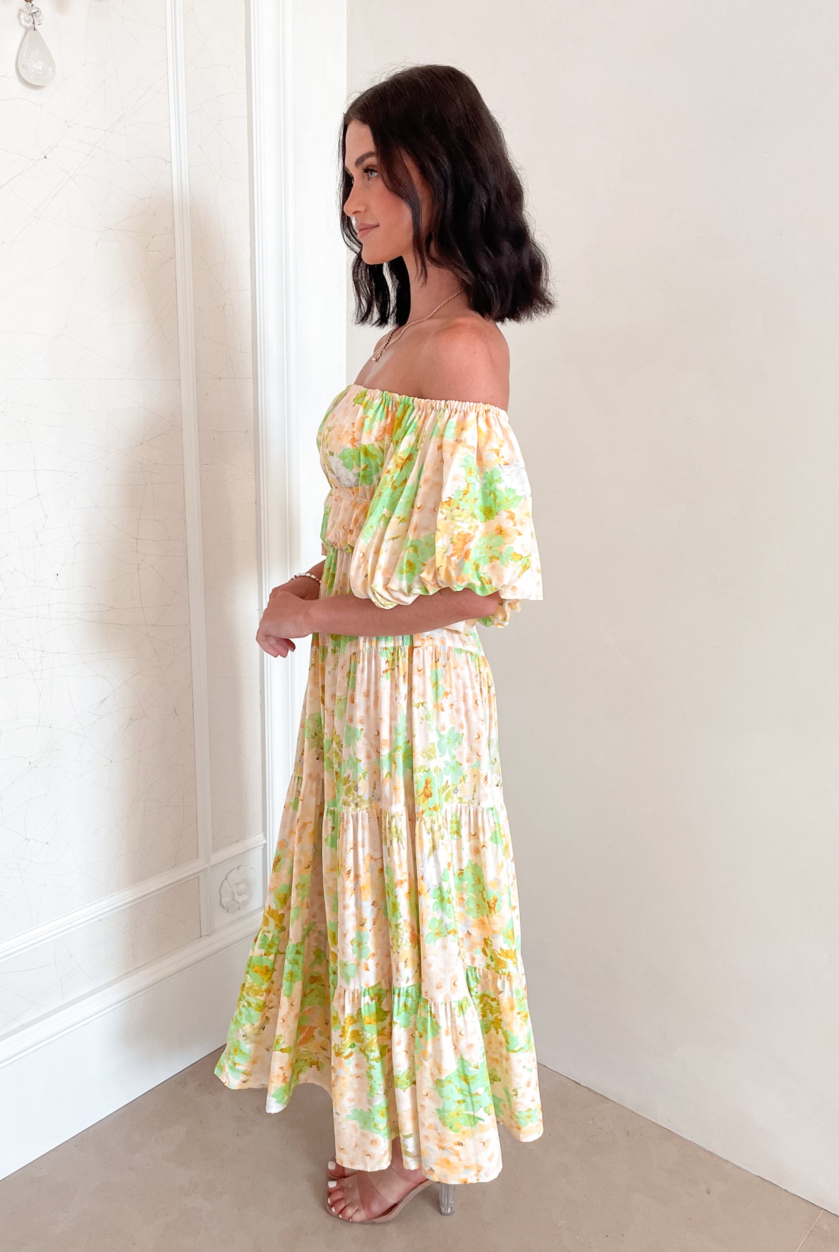 A woman with shoulder-length dark hair stands against a light beige wall, wearing an off-the-shoulder, flowy midi dress with a pastel floral pattern in shades of yellow, green, and peach. The dress features puffed sleeves and a smocked waist. She completes the look with clear strap heels and a delicate necklace, posing gracefully with her hands gently clasped in front.