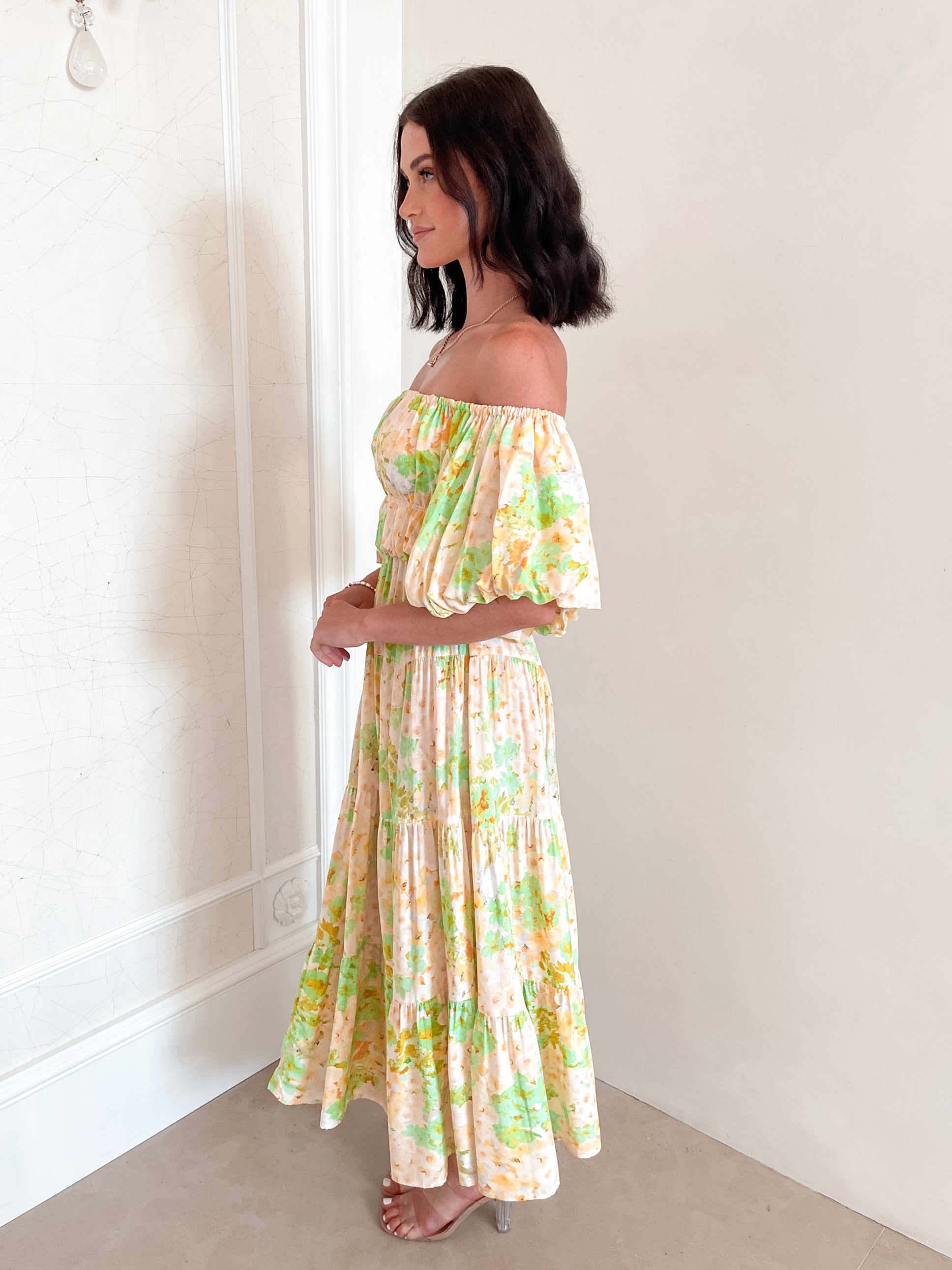 A woman with shoulder-length dark hair stands against a light beige wall, wearing an off-the-shoulder, flowy midi dress with a pastel floral pattern in shades of yellow, green, and peach. The dress features puffed sleeves and a smocked waist. She completes the look with clear strap heels and a delicate necklace, posing gracefully with her hands gently clasped in front.