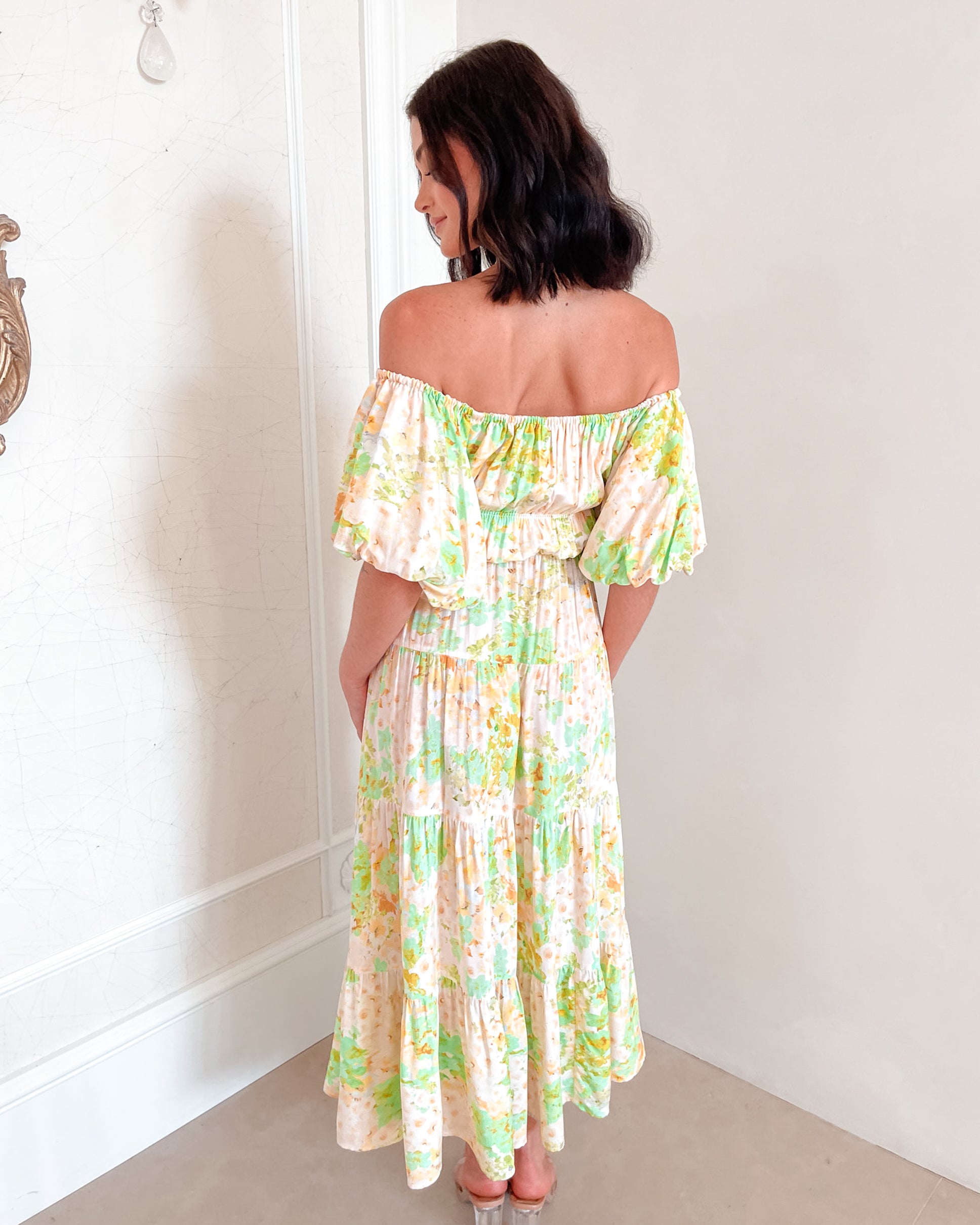 A woman with shoulder-length dark hair stands against a light beige wall, wearing an off-the-shoulder, flowy midi dress with a pastel floral pattern in shades of yellow, green, and peach. The dress features puffed sleeves and a smocked waist. She completes the look with clear strap heels and a delicate necklace, posing gracefully with her hands gently clasped in front.