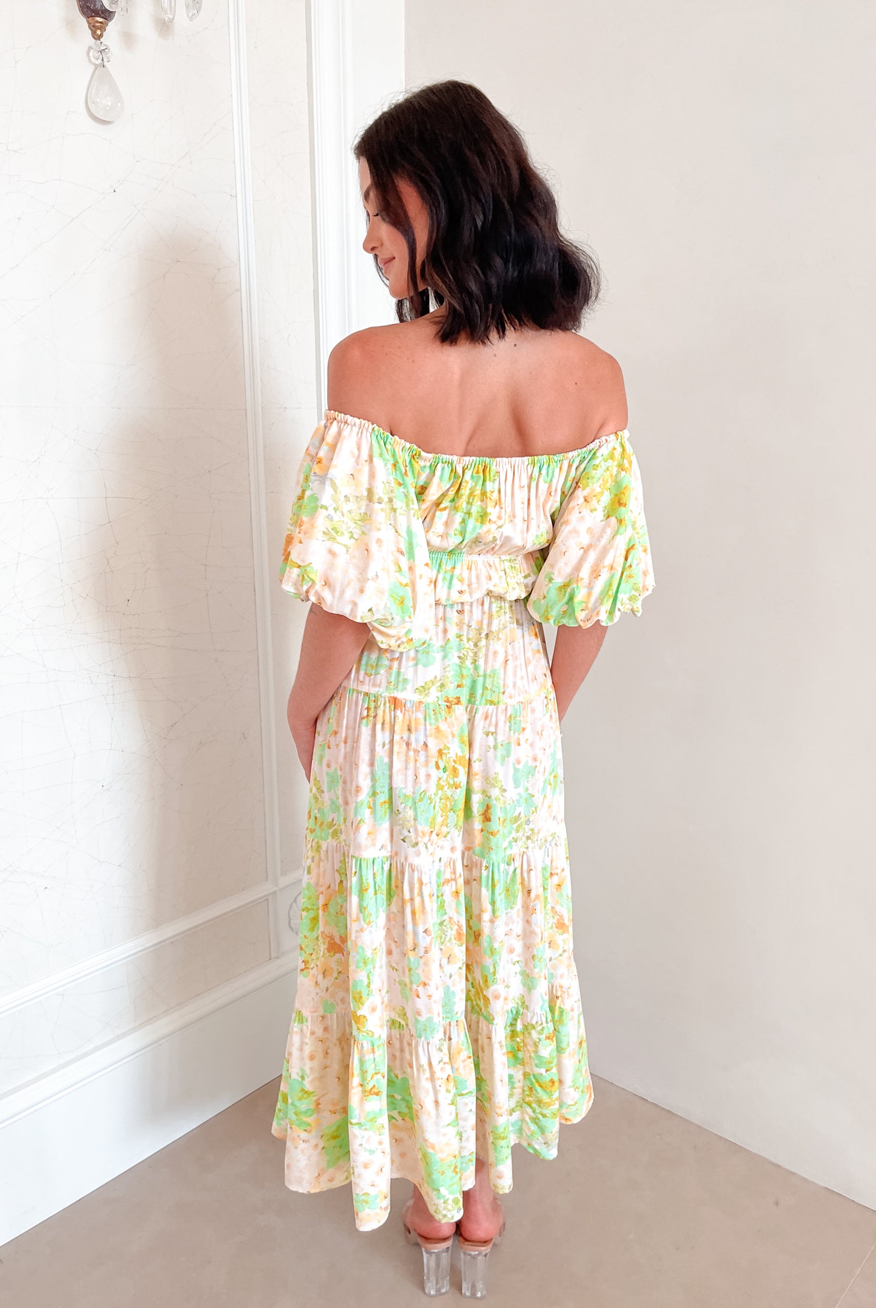 A woman with shoulder-length dark hair stands against a light beige wall, wearing an off-the-shoulder, flowy midi dress with a pastel floral pattern in shades of yellow, green, and peach. The dress features puffed sleeves and a smocked waist. She completes the look with clear strap heels and a delicate necklace, posing gracefully with her hands gently clasped in front.