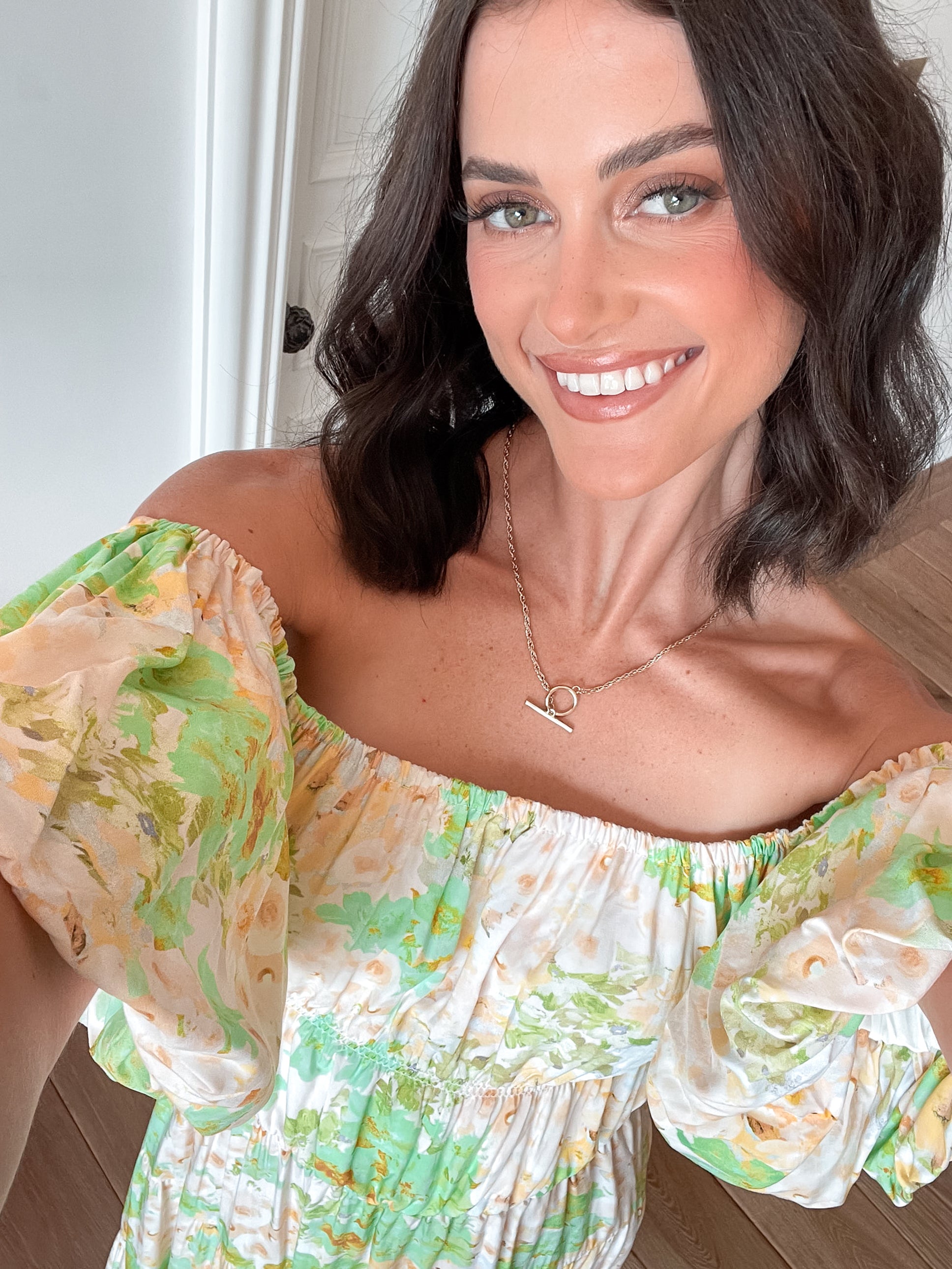 A woman with shoulder-length dark hair stands against a light beige wall, wearing an off-the-shoulder, flowy midi dress with a pastel floral pattern in shades of yellow, green, and peach. The dress features puffed sleeves and a smocked waist. She completes the look with clear strap heels and a delicate necklace, posing gracefully with her hands gently clasped in front.