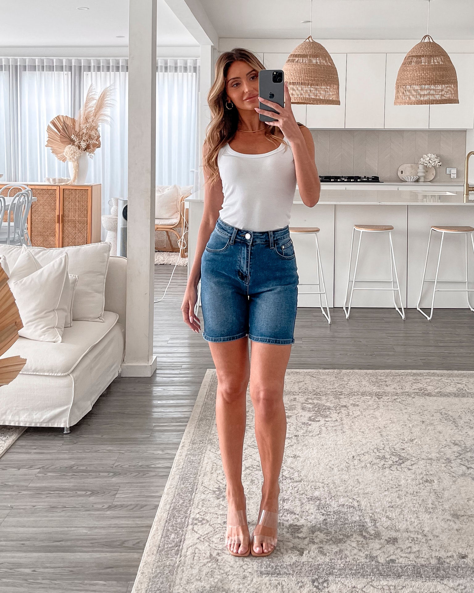 A smiling woman with long, wavy hair stands indoors in front of a white distressed wooden sideboard decorated with a large shell sculpture, a white vase holding dried palm leaves, and a round wall clock. She is wearing a white sleeveless tank top paired with high-waisted denim shorts and clear strap heels, with one hand resting in her pocket. The setting has a cozy, neutral-toned decor with a woven basket to the side and a patterned rug underfoot.