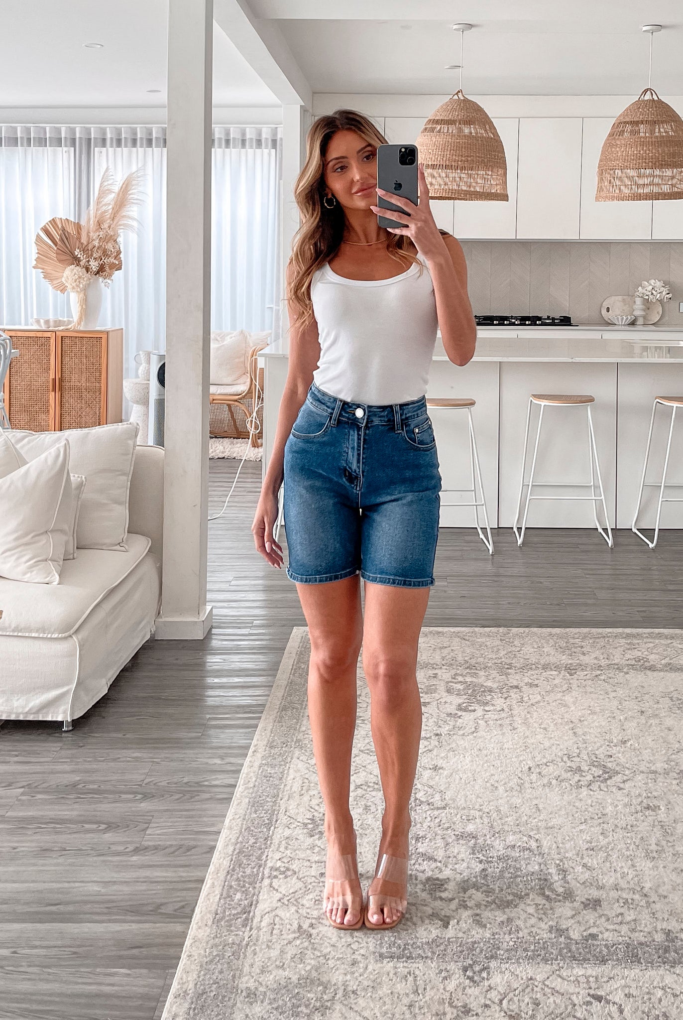 A smiling woman with long, wavy hair stands indoors in front of a white distressed wooden sideboard decorated with a large shell sculpture, a white vase holding dried palm leaves, and a round wall clock. She is wearing a white sleeveless tank top paired with high-waisted denim shorts and clear strap heels, with one hand resting in her pocket. The setting has a cozy, neutral-toned decor with a woven basket to the side and a patterned rug underfoot.