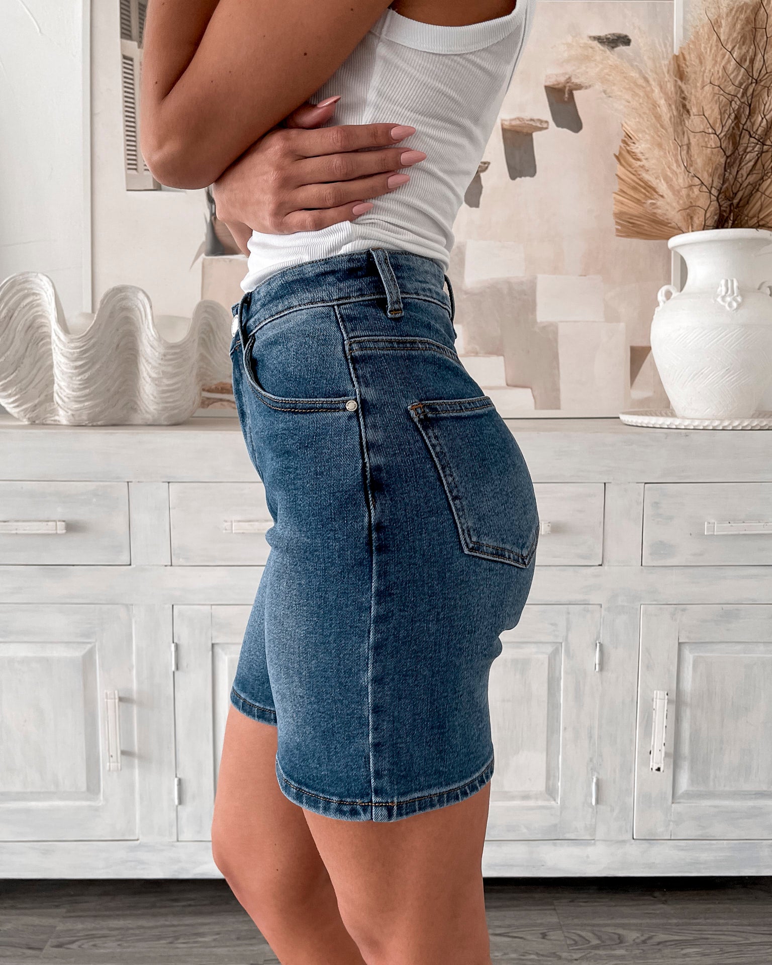 A smiling woman with long, wavy hair stands indoors in front of a white distressed wooden sideboard decorated with a large shell sculpture, a white vase holding dried palm leaves, and a round wall clock. She is wearing a white sleeveless tank top paired with high-waisted denim shorts and clear strap heels, with one hand resting in her pocket. The setting has a cozy, neutral-toned decor with a woven basket to the side and a patterned rug underfoot.