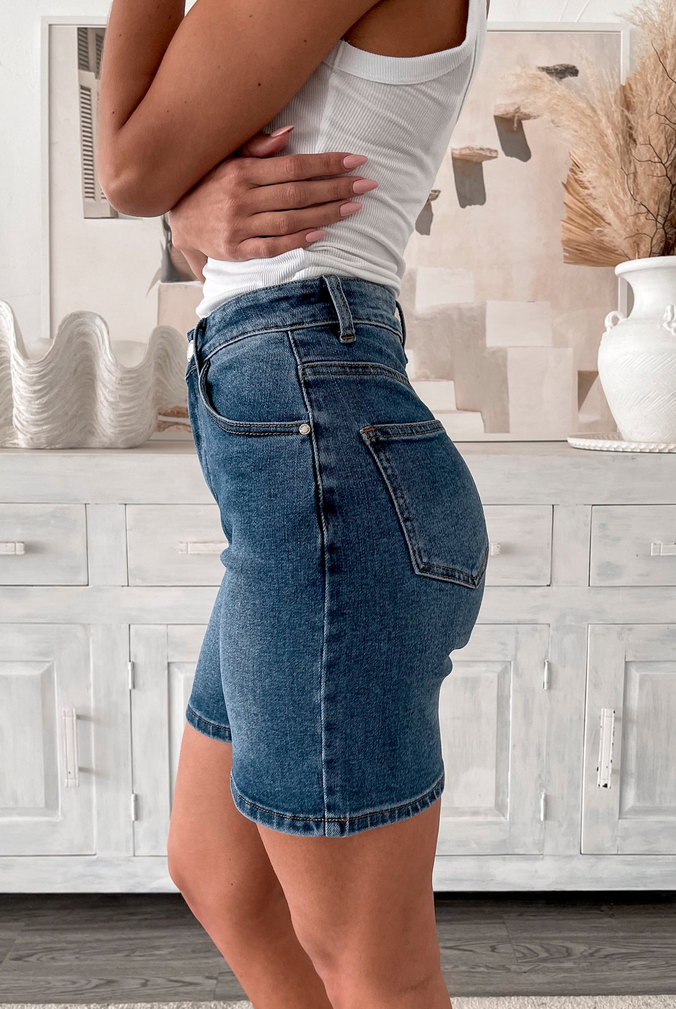 A smiling woman with long, wavy hair stands indoors in front of a white distressed wooden sideboard decorated with a large shell sculpture, a white vase holding dried palm leaves, and a round wall clock. She is wearing a white sleeveless tank top paired with high-waisted denim shorts and clear strap heels, with one hand resting in her pocket. The setting has a cozy, neutral-toned decor with a woven basket to the side and a patterned rug underfoot.