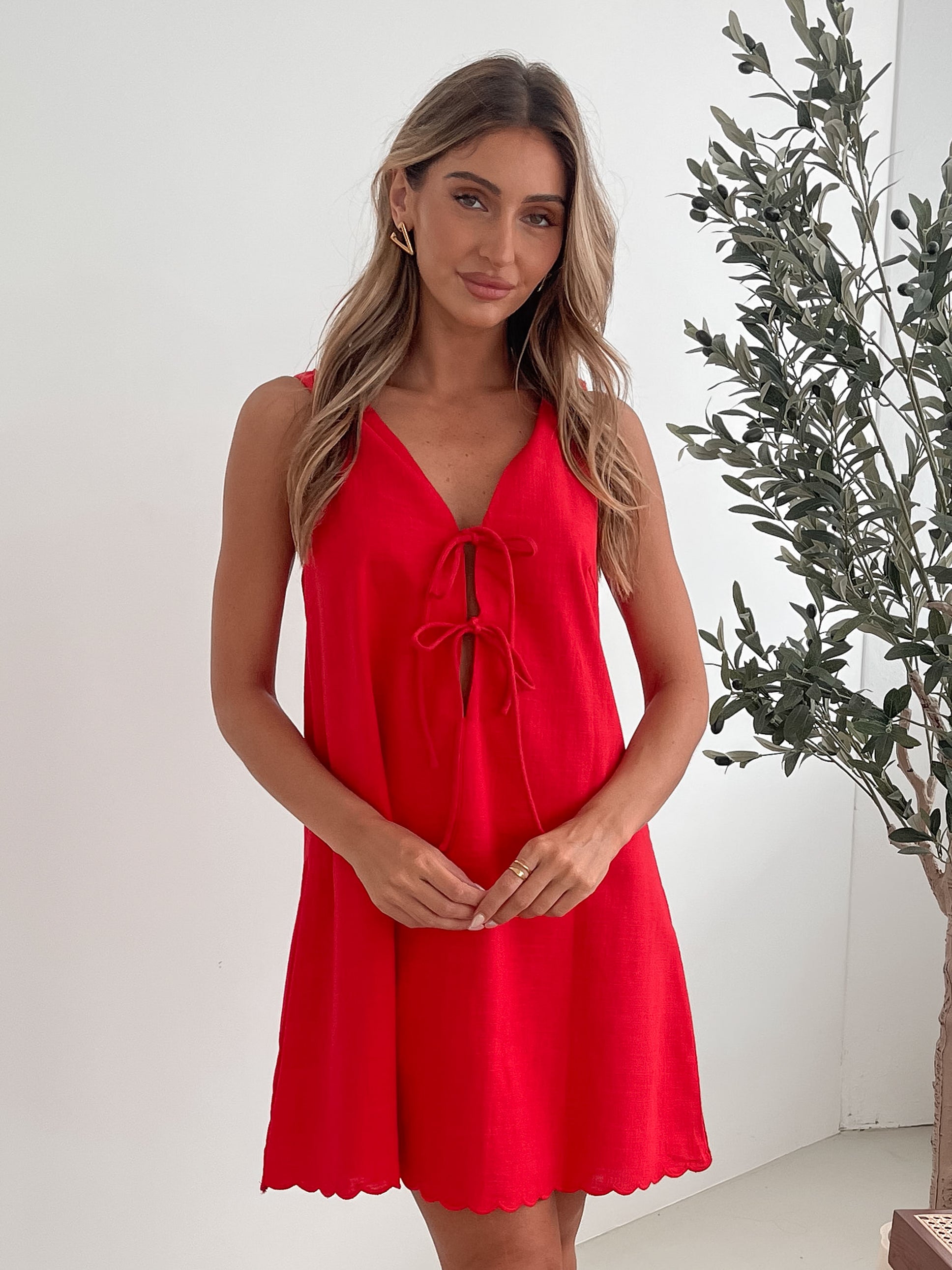 A woman with long, wavy blonde hair stands indoors in front of a plain white wall, with a green leafy plant beside her. She is wearing a sleeveless red dress with a V-neck and two front tie details, similar in style to the previous dress, featuring a scalloped hem. Her hands are gently clasped together in front of her, and she has a subtle smile on her face. She accessorizes with gold hoop earrings and a delicate ring, and her makeup is natural and polished.